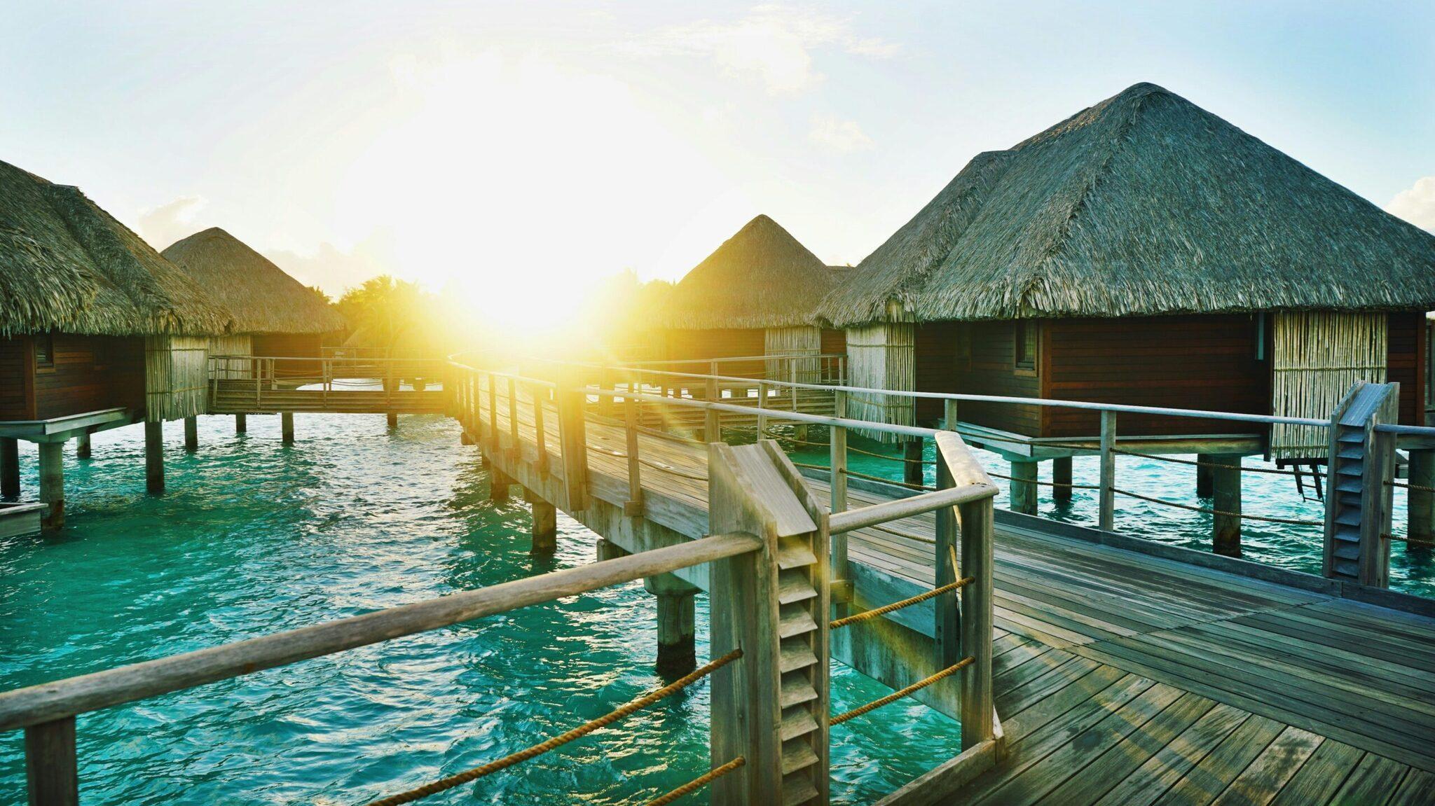 Bungalows sur pilotis avec toits de chaume surplombant une lagune turquoise au coucher de soleil