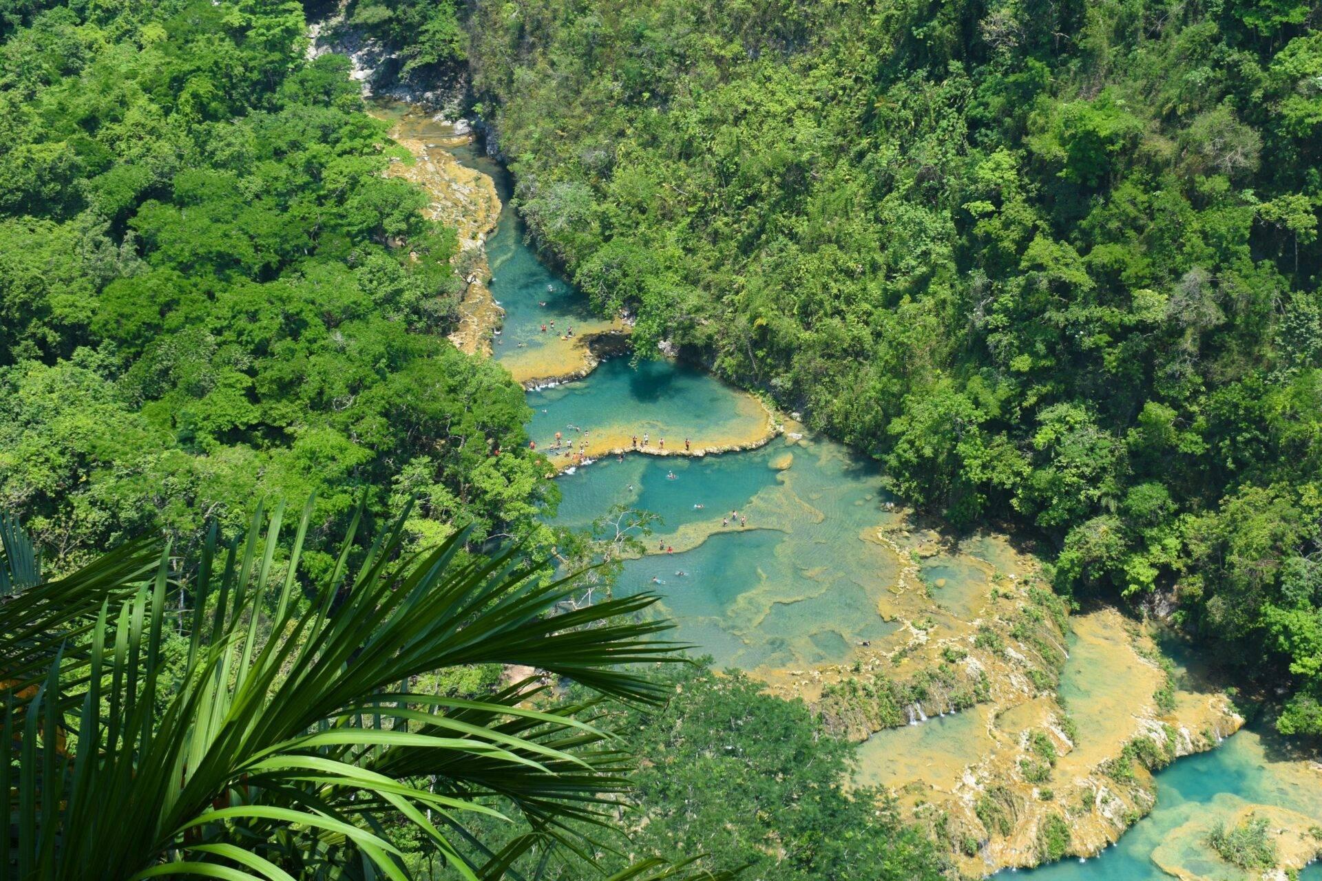 Vue aérienne d’une rivière turquoise serpentant dans une jungle dense avec baigneurs, entourée de falaises calcaires