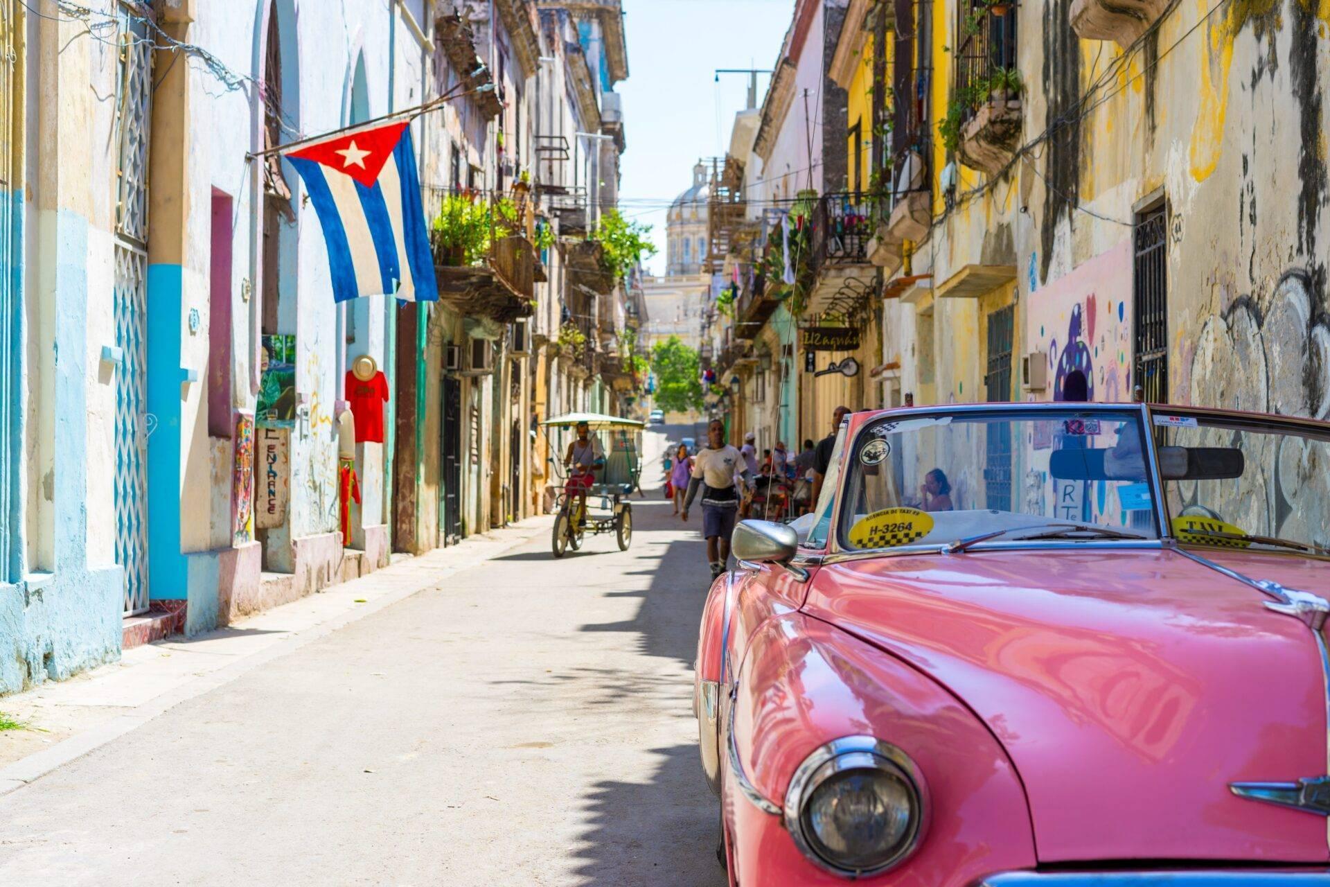 Une rue pavée colorée avec une voiture vintage rouge, un drapeau cubain et des bâtiments historiques aux façades délabrées