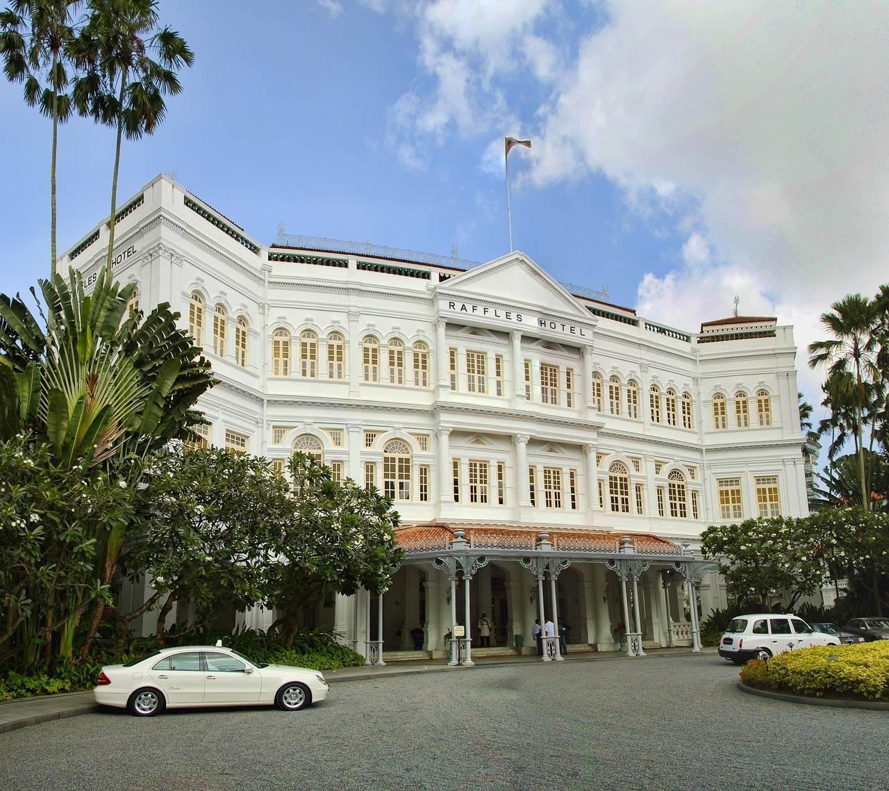 Un hôtel colonial blanc majestueux avec colonnes, entouré de palmiers tropicaux et de voitures garées devant