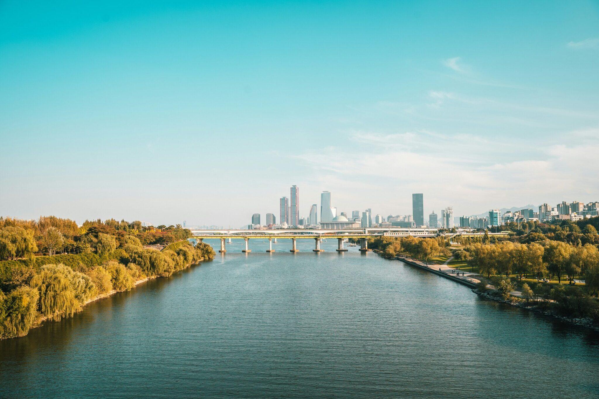 View of a river with a bridge and modern skyscrapers in the background, lined with golden trees in autumn