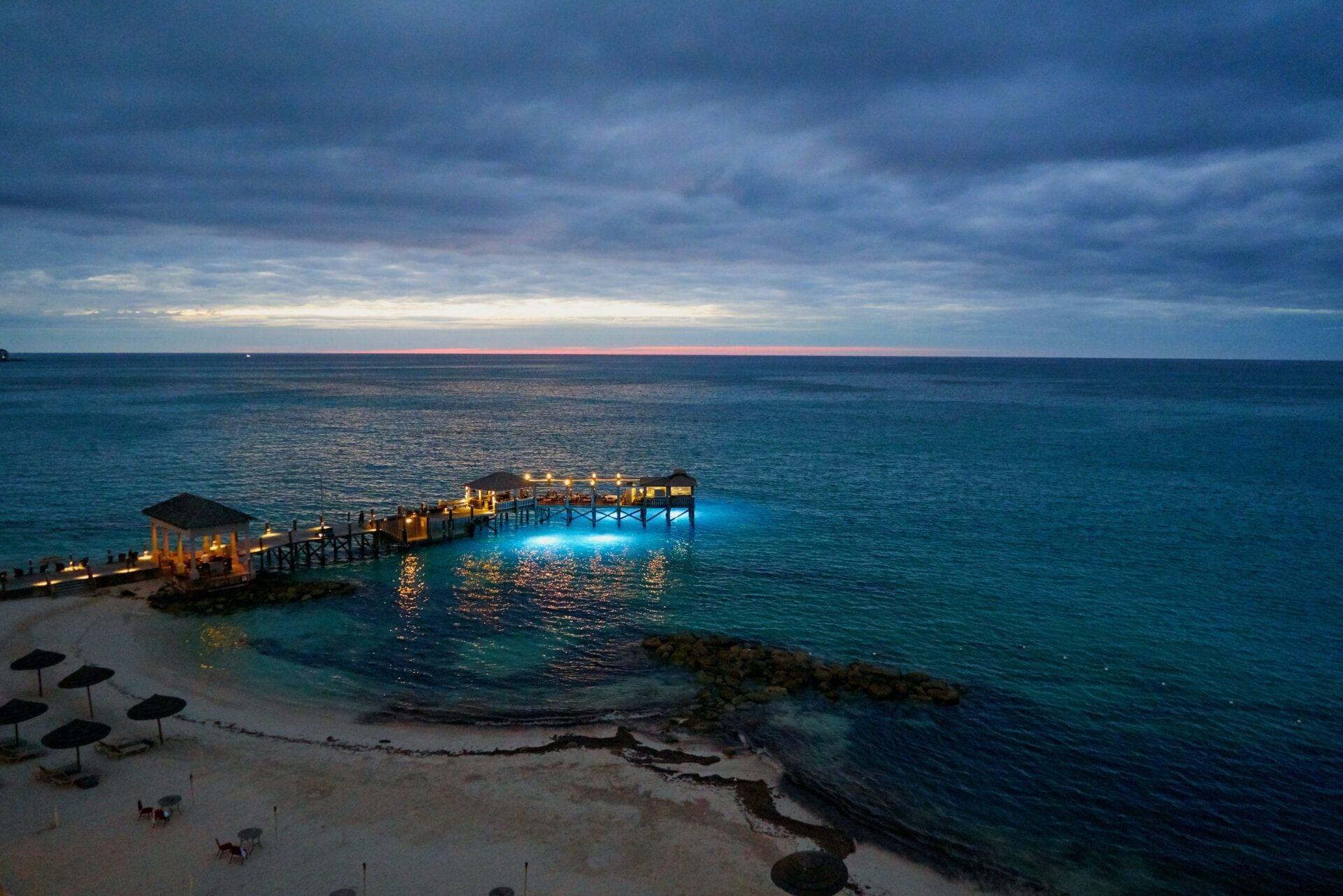 Un ponton illuminé s’étendant dans une mer turquoise au crépuscule, avec plage et parasols
