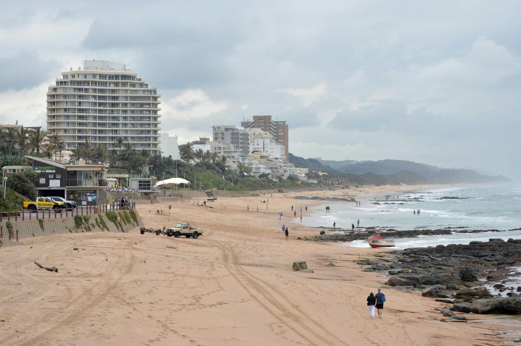 A coastal beach with residential buildings, black rocks, swimmers and vehicles on the sand