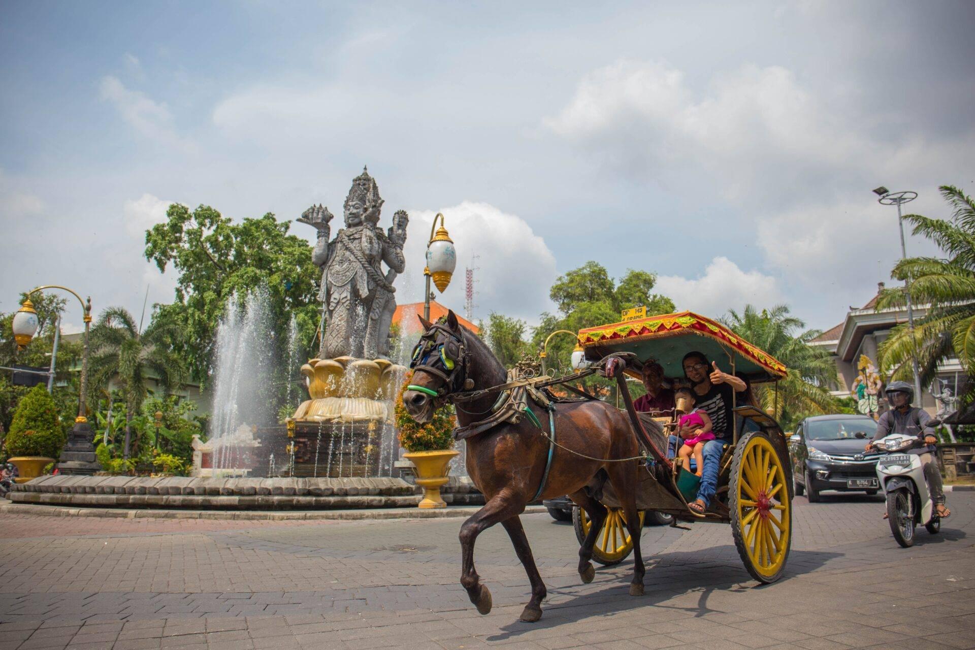 A traditional yellow and red horse-drawn float passes a divine statue in an Indonesian public square