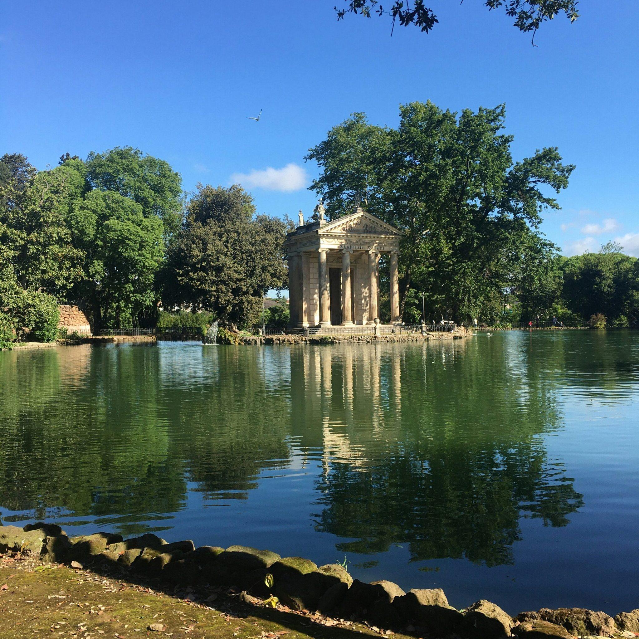 A classical columned temple reflected in a calm lake, surrounded by lush greenery in fine weather