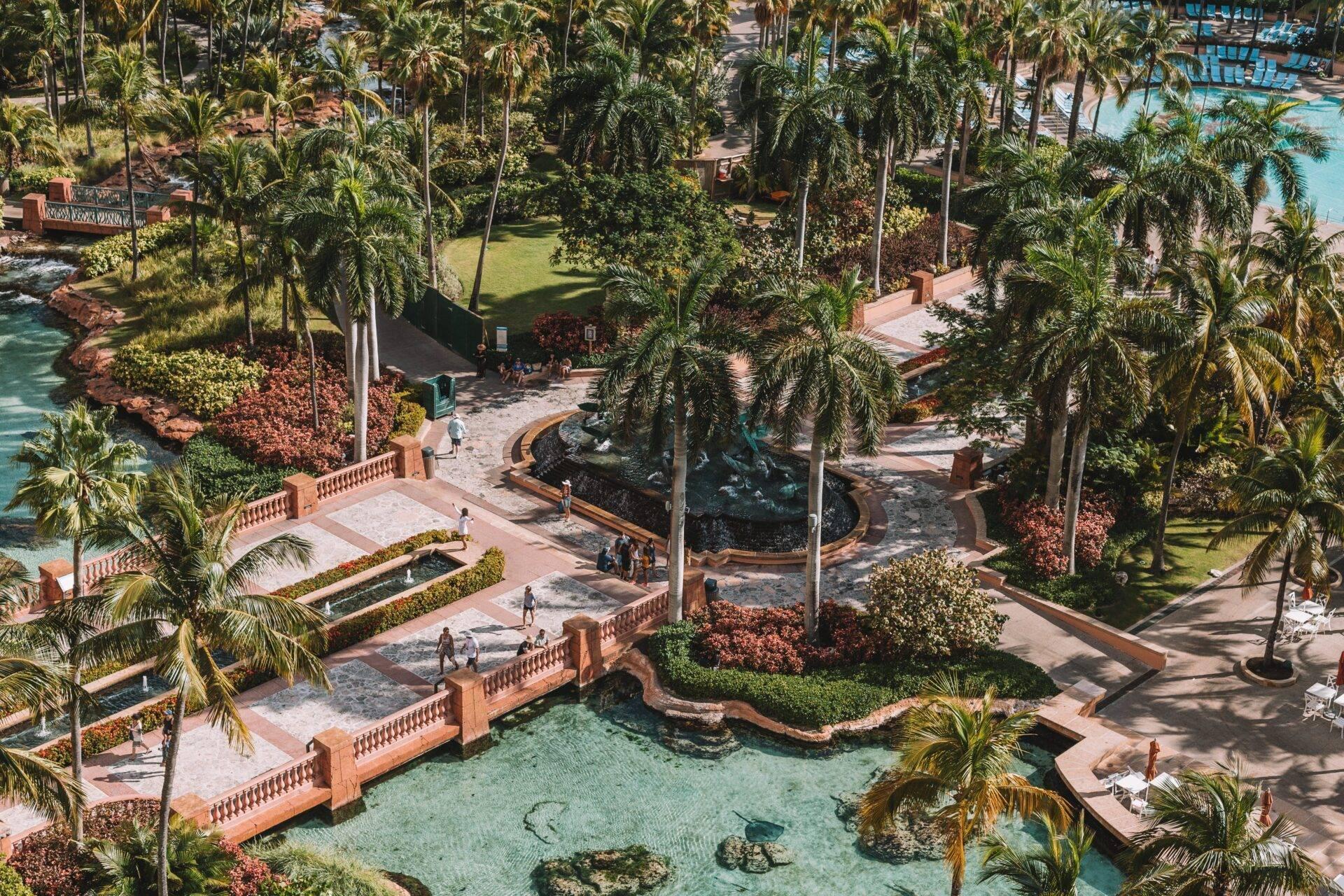 Vue aérienne d’un resort tropical avec bassins d’eau turquoise, terrasses en brique, palmiers et sculptures, entouré de verdure