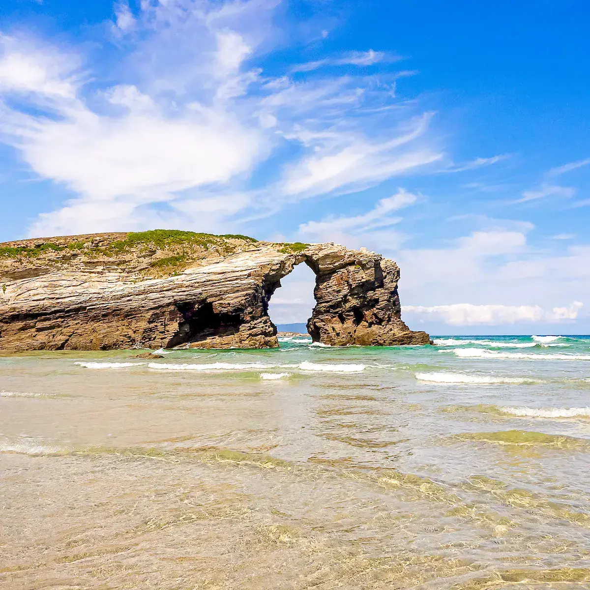 Une arche rocheuse naturelle émergeant de l’océan sur une plage de sable fin sous un ciel bleu dégagé