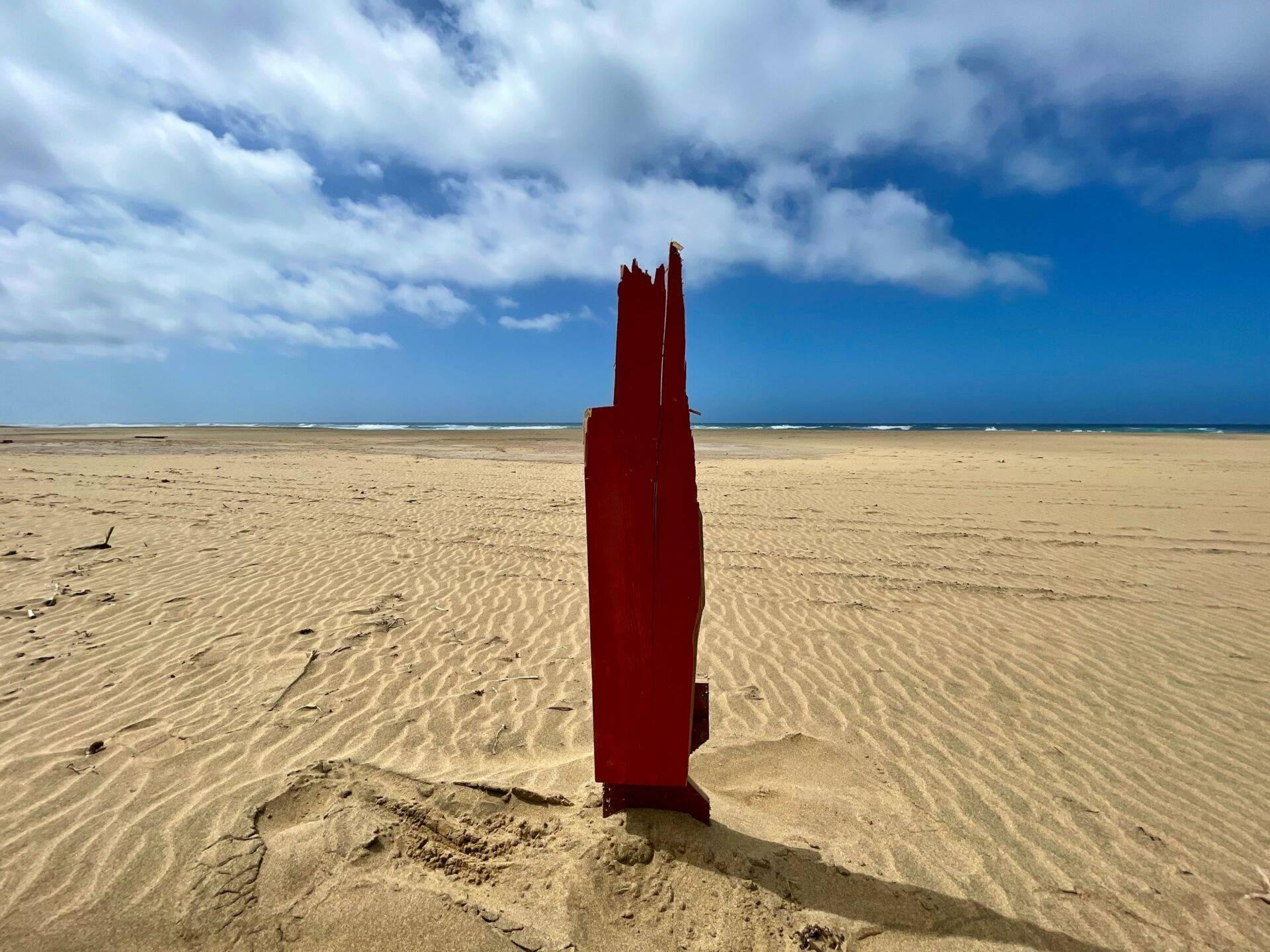 Une structure métallique rouge rouillée émerge du sable d’une vaste plage déserte aux Canaries sous un ciel nuageux