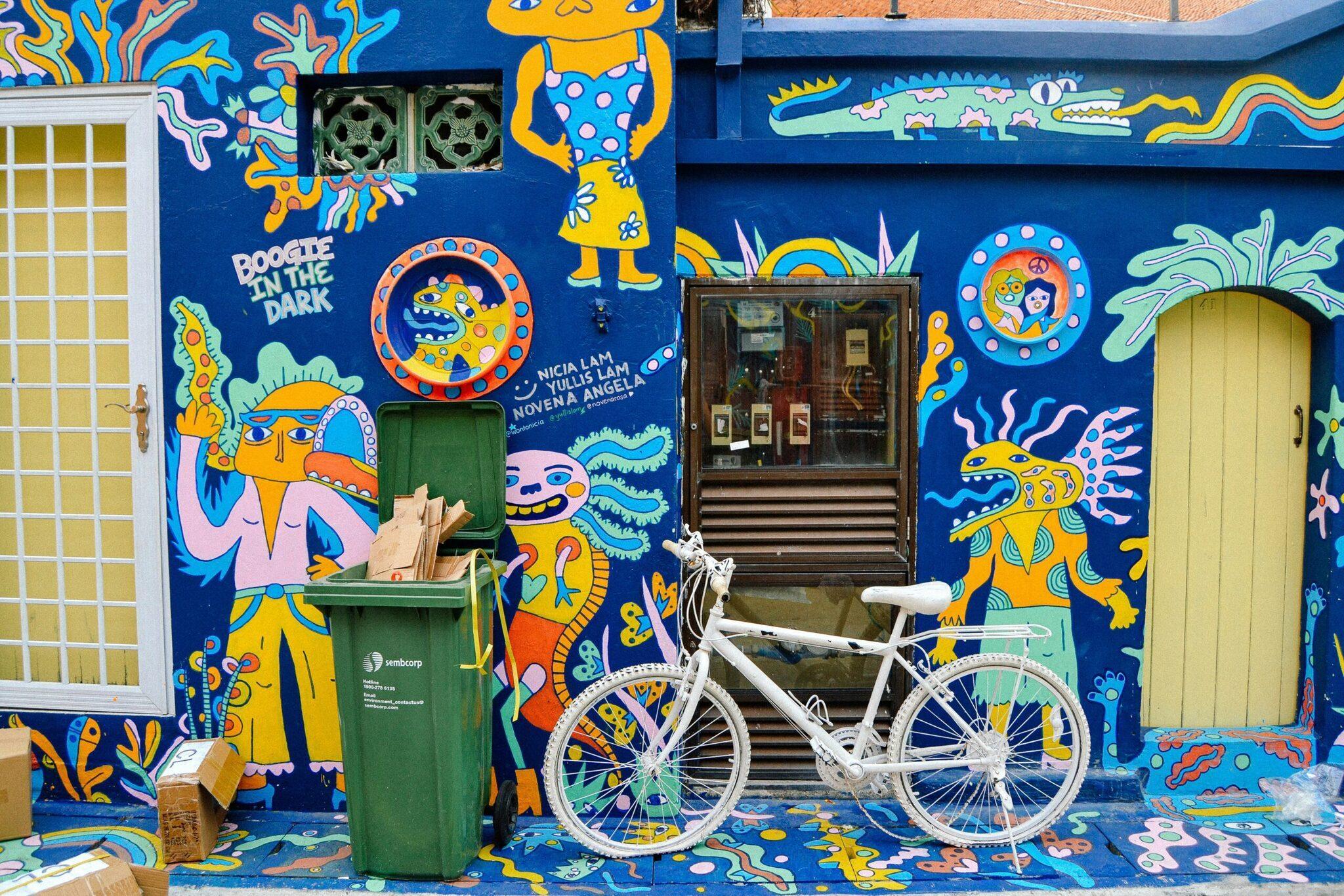 Un vélo blanc devant une façade bleue ornée de fresques colorées et de motifs tropicaux stylisés