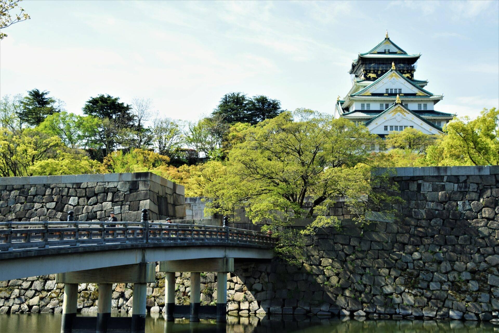 A traditional Japanese castle with curved rooflines dominating a green landscape crossed by a stone bridge