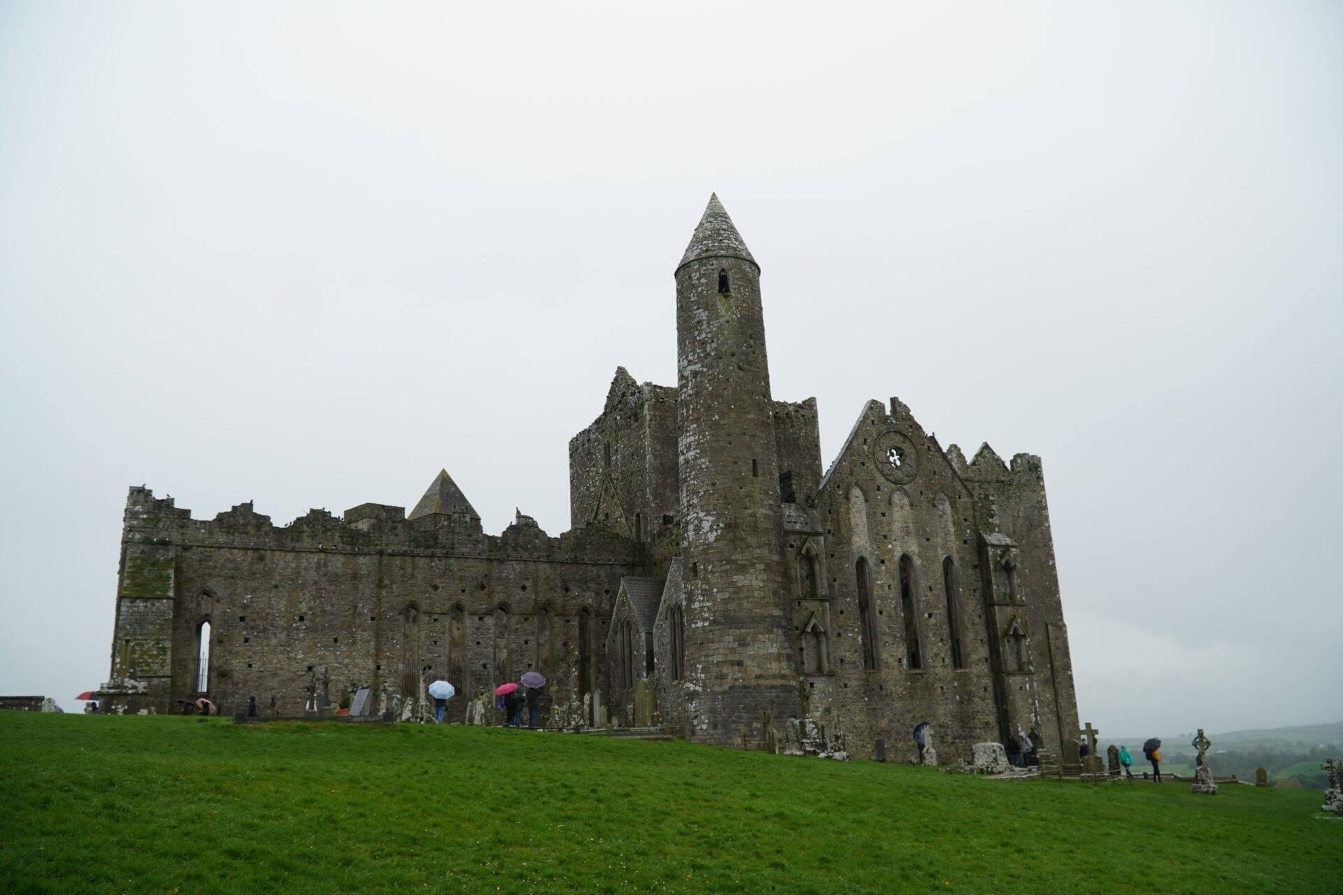 Ruins of a stone medieval abbey with a round tower and graveyard under a grey cloudy sky