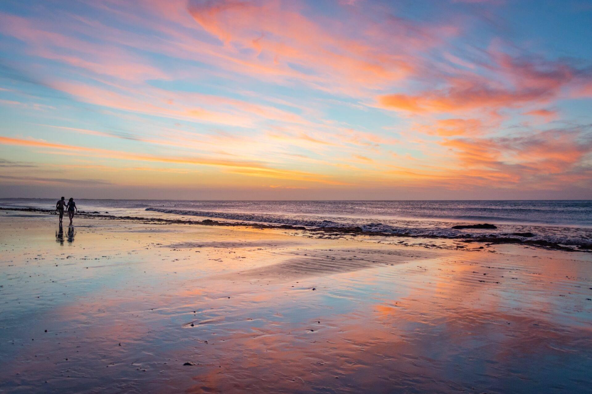 Two surfers on a beach at sunset with golden reflections shimmering on the wet sand