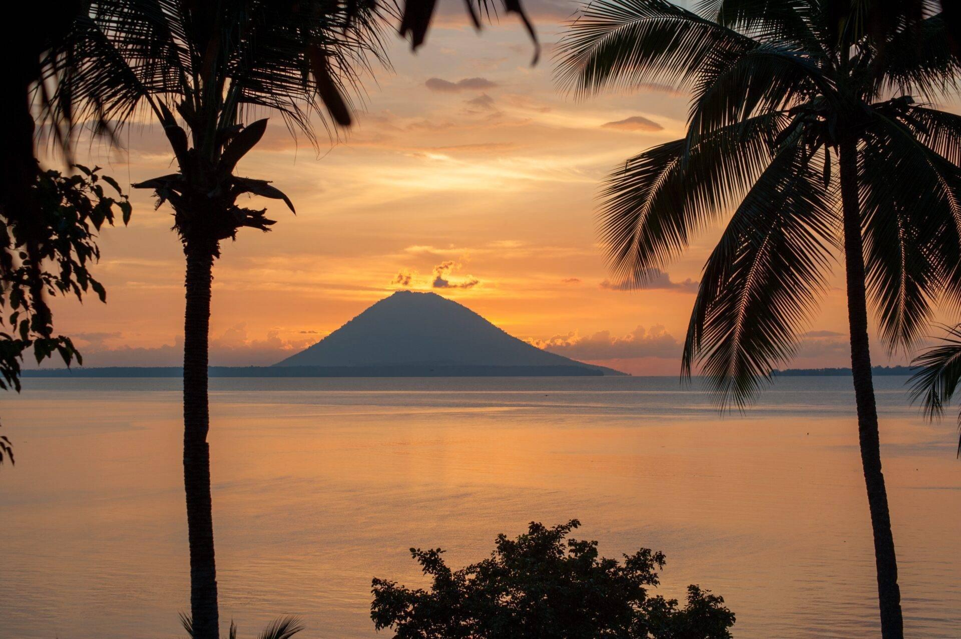 A tropical sunset over a volcano rising from the sea, framed by silhouetted palm trees