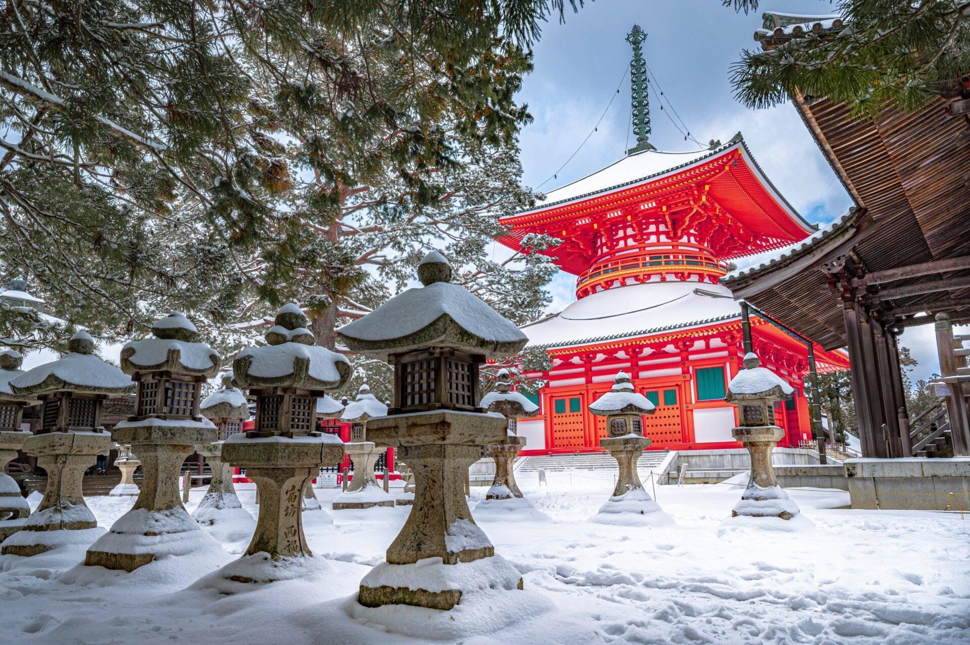 A Japanese Buddhist temple with vivid red rooftops surrounded by snow-dusted stone lanterns beneath frost-covered trees