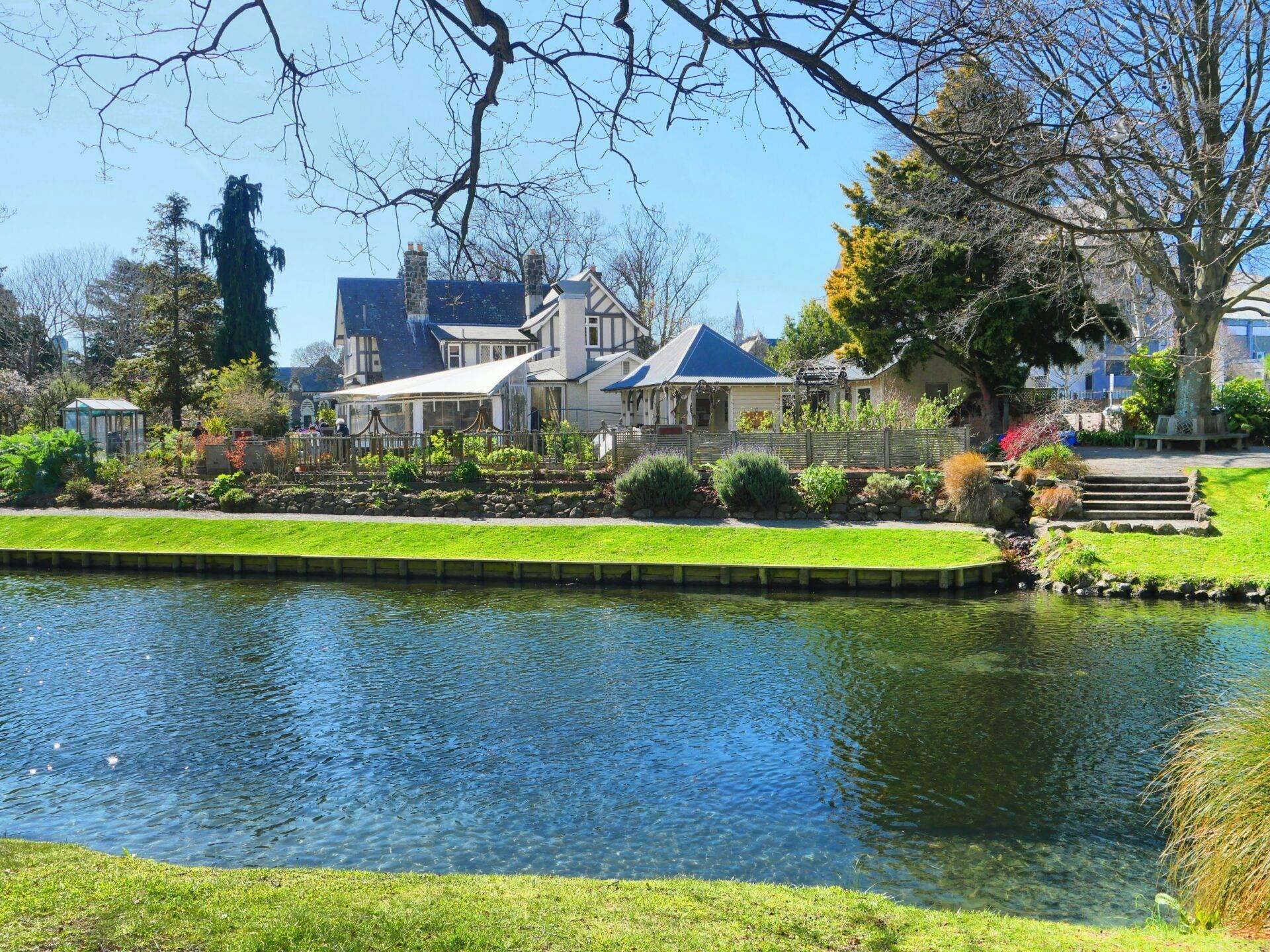 Une maison blanche au bord d’une rivière avec jardin fleuri et arbres nus, paysage paisible de campagne verdoyante