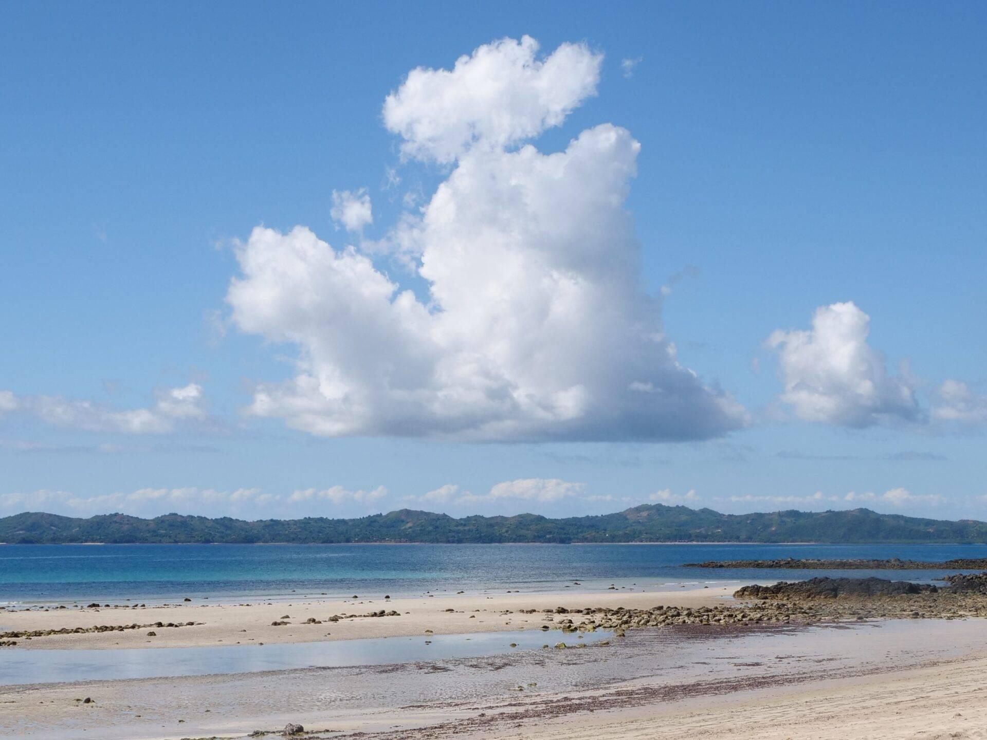 White sand beach with a turquoise lagoon, wooded mountains in the background and a partly cloudy blue sky
