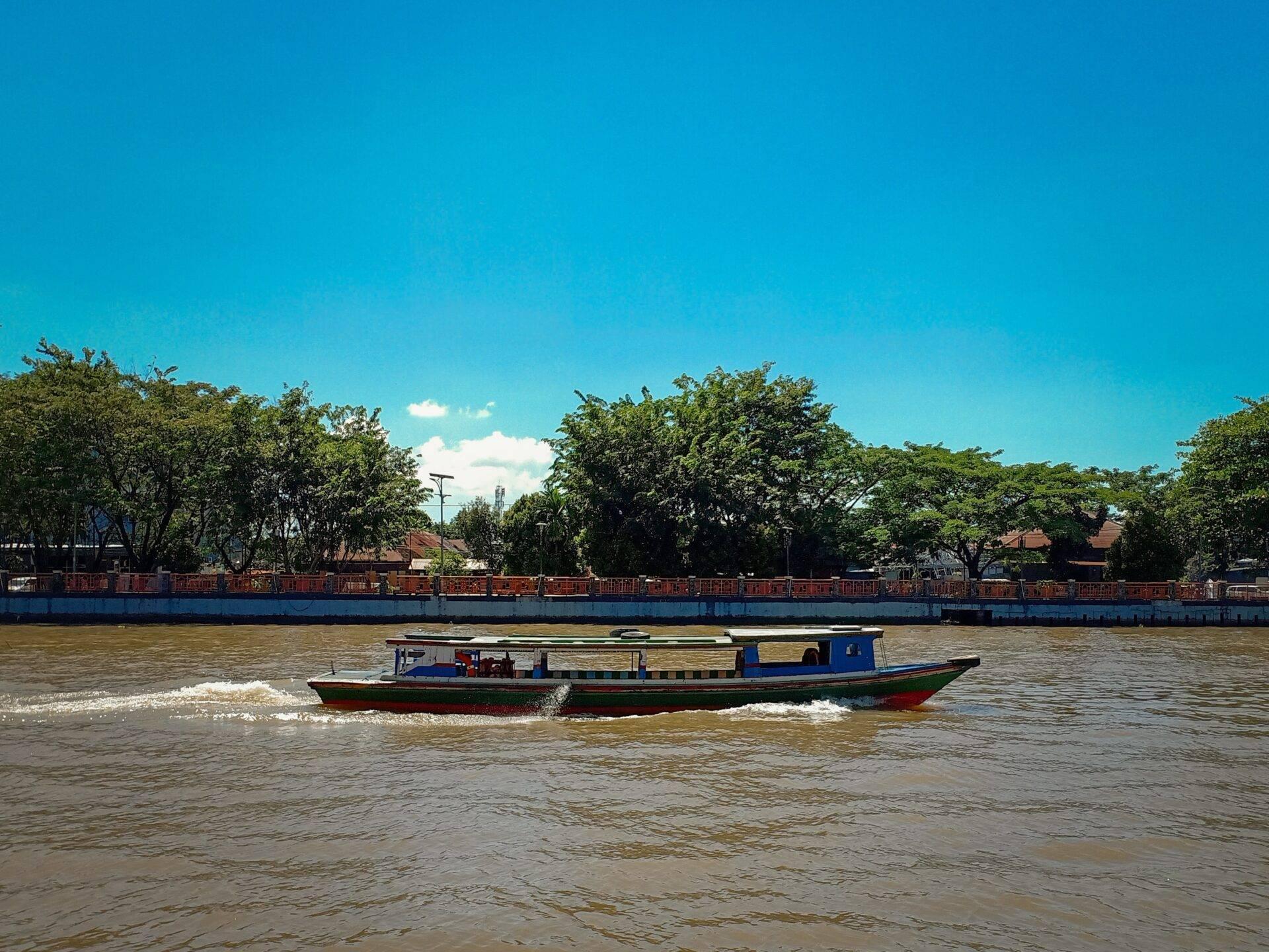 A colourful river transport boat navigates a brown river under a clear blue sky, with trees and port structures in the background