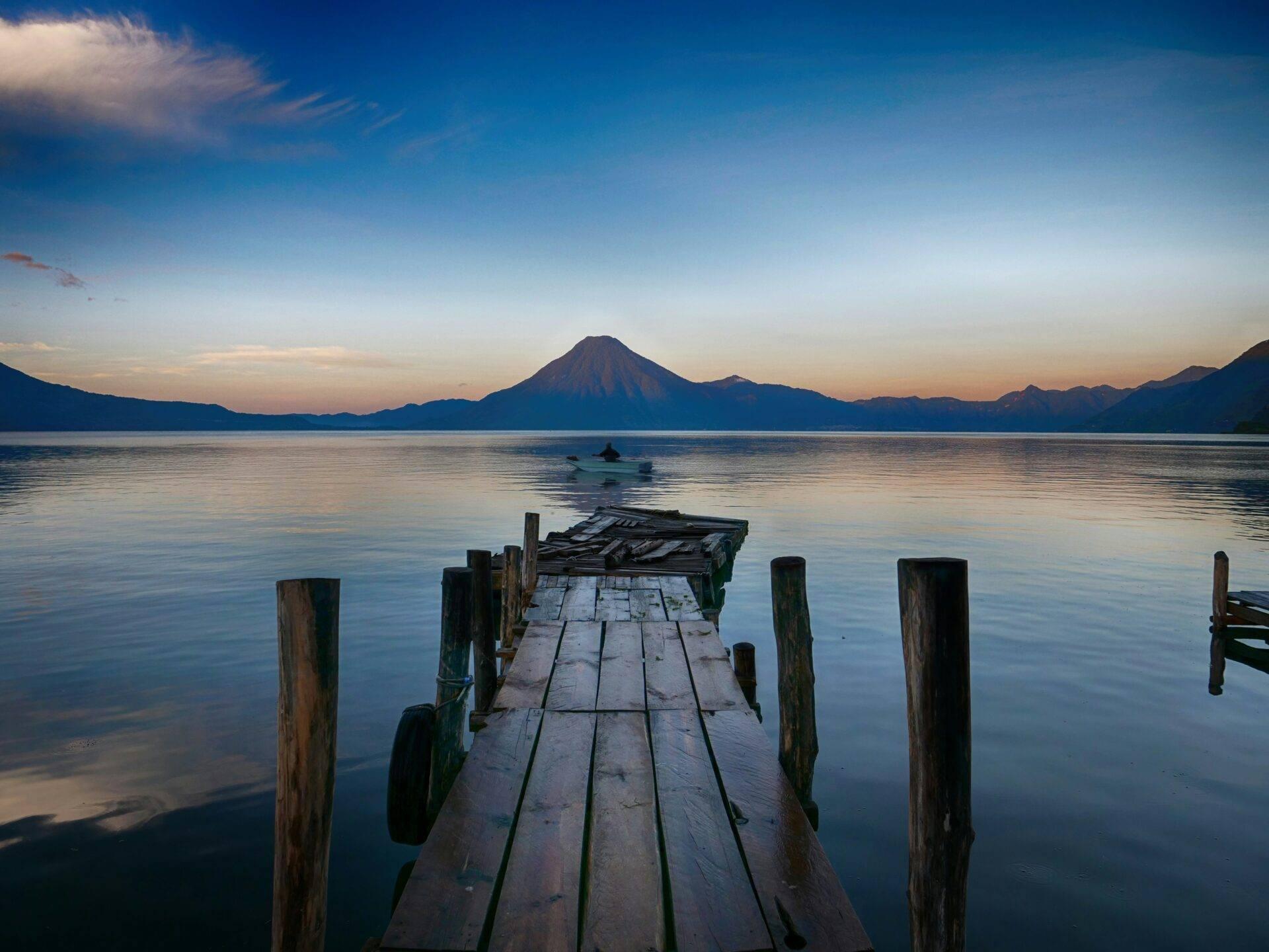 Un ponton en bois s’avance dans un lac calme avec un volcan majestueux en arrière-plan au crépuscule