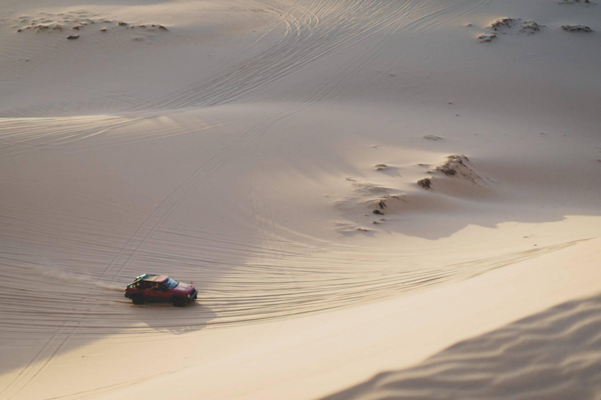 Un véhicule rouge traverse une vaste plage de sable blanc aux motifs ondulés sous une lumière douce