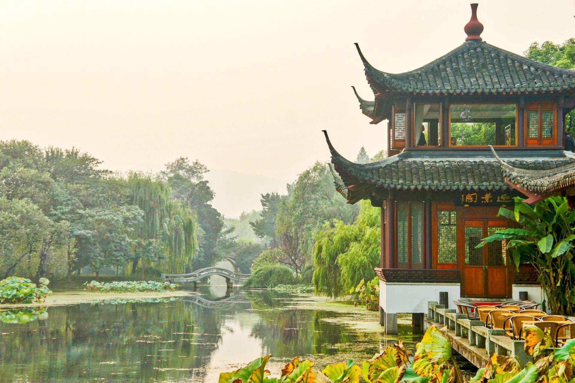 Pavillon traditionnel chinois aux toits recourbés surplombant un canal avec pont arqué et saules pleureurs