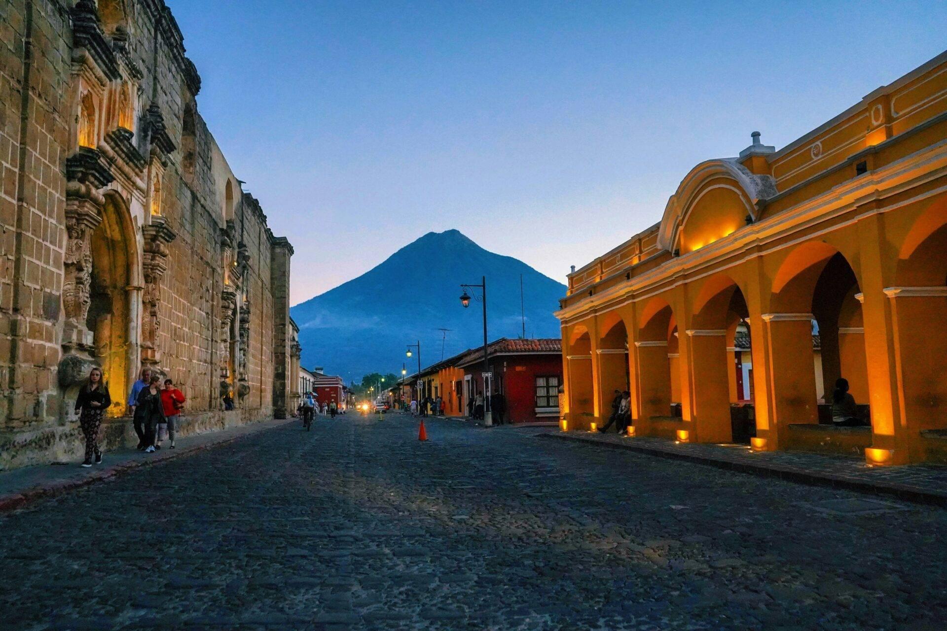 Rue coloniale pavée avec arcades jaunes illuminées et volcan en arrière-plan au crépuscule