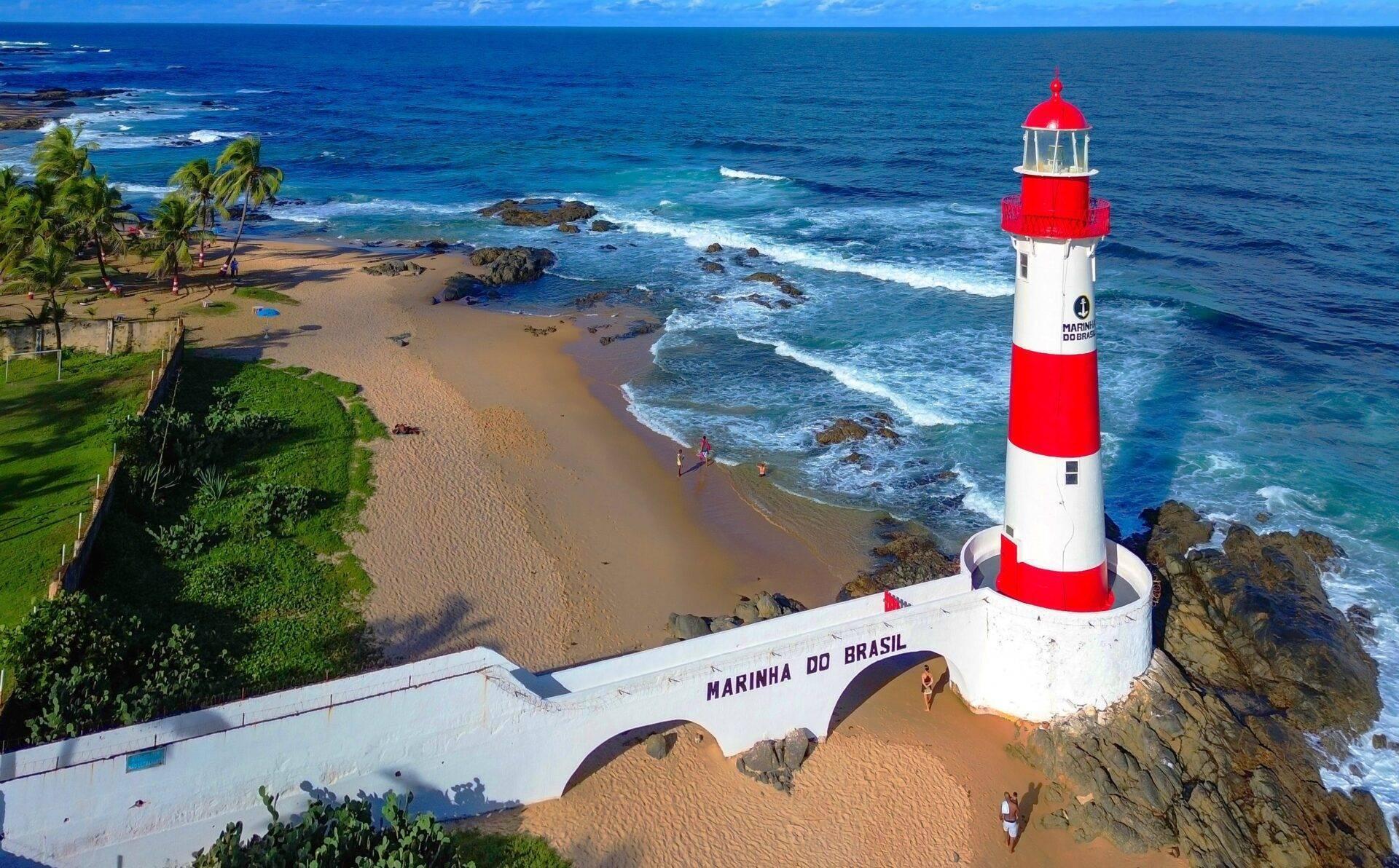 Phare rouge et blanc en bord de plage avec palmiers, rochers et océan bleu sous ciel dégagé au Brésil