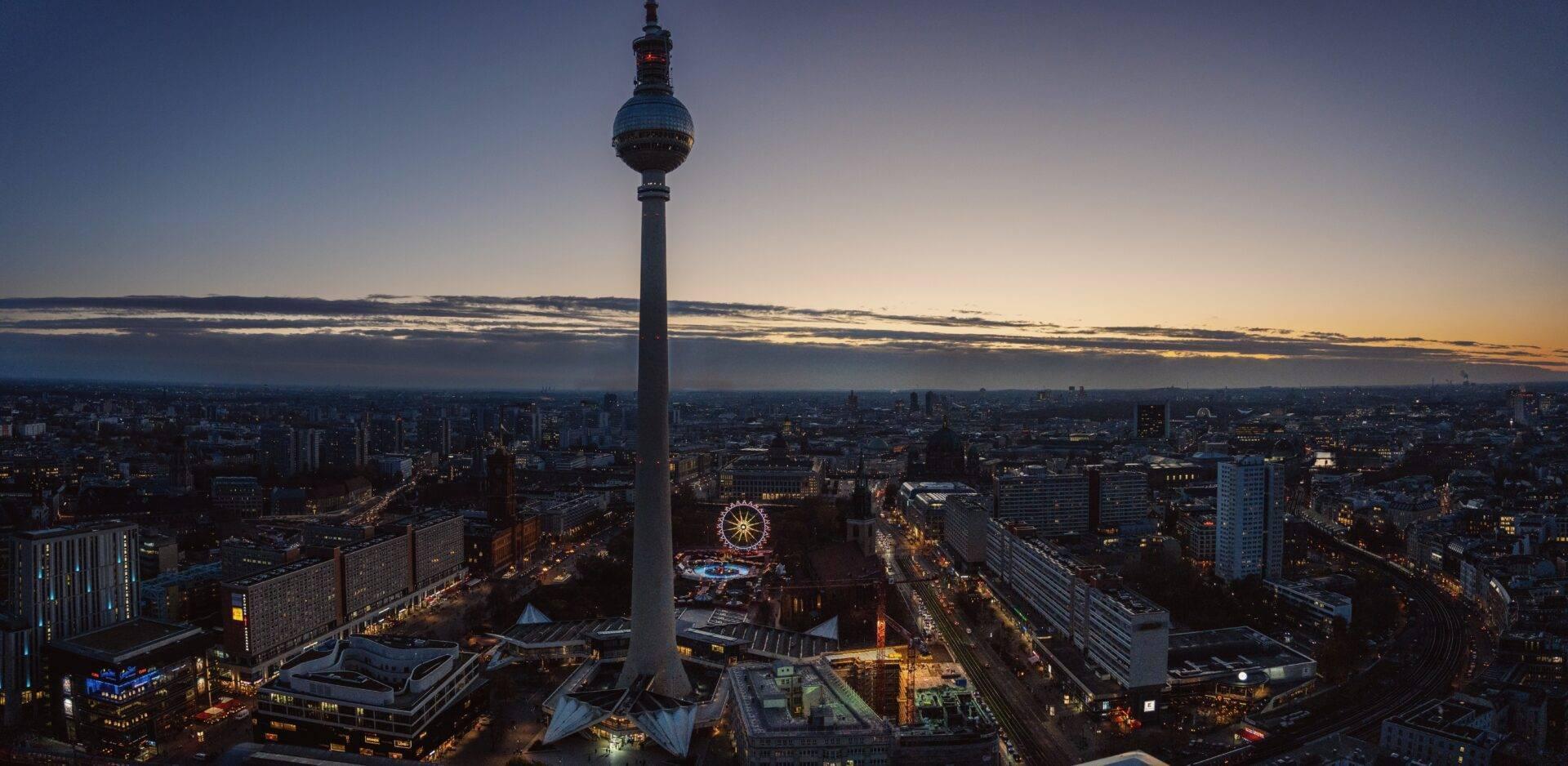 Panoramic dusk view of a tall television tower dominating a German city skyline, with an illuminated Ferris wheel