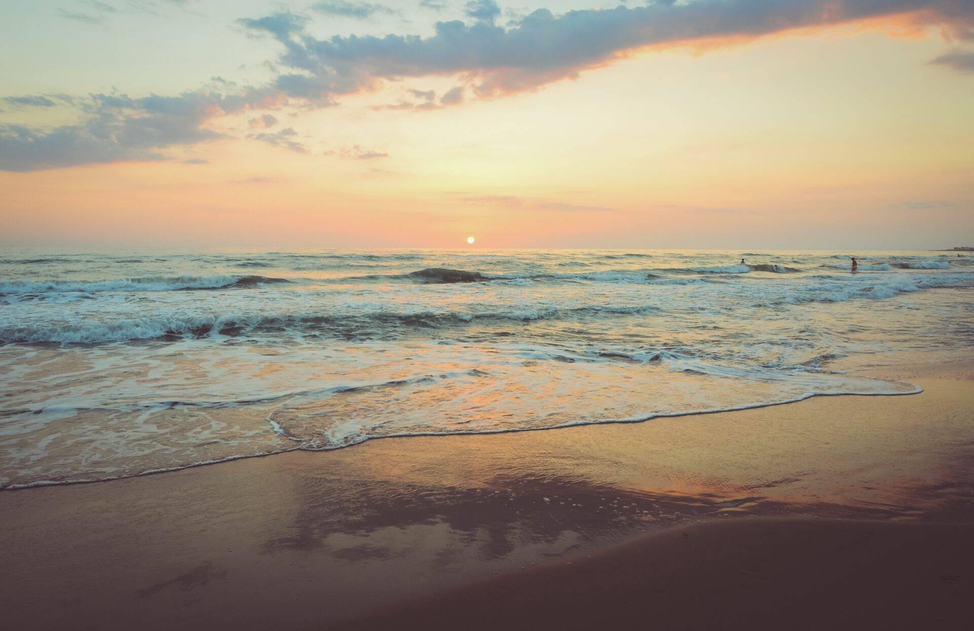 Sunset over a sandy beach with waves rolling in and a handful of swimmers visible on the horizon