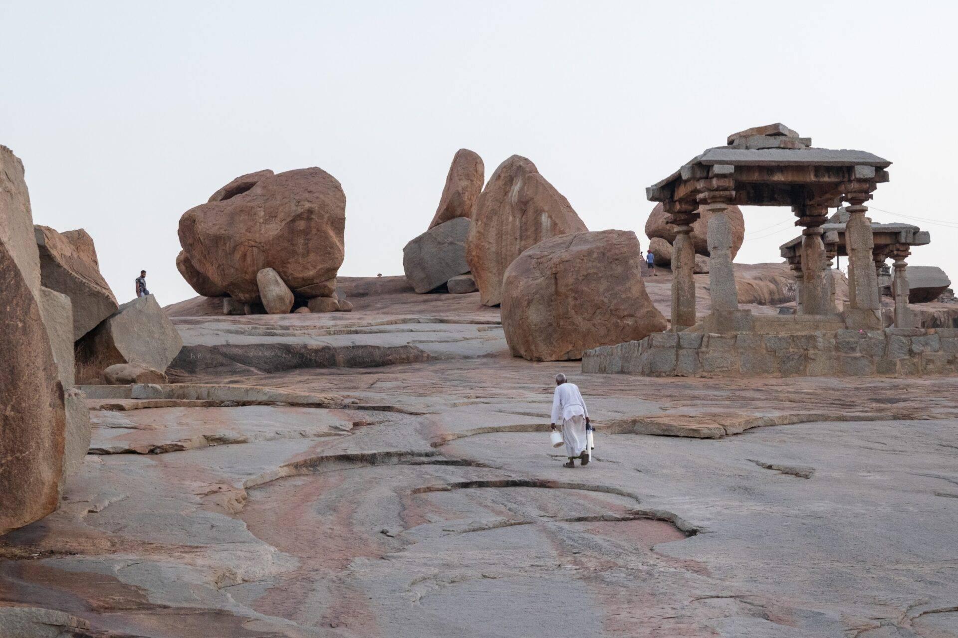 A man in white clothing walks across a rocky plateau scattered with giant boulders and stone temple ruins