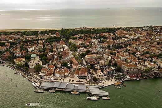 Aerial view of a European coastal city with dense historic architecture, a picturesque waterfront and a maritime vessel