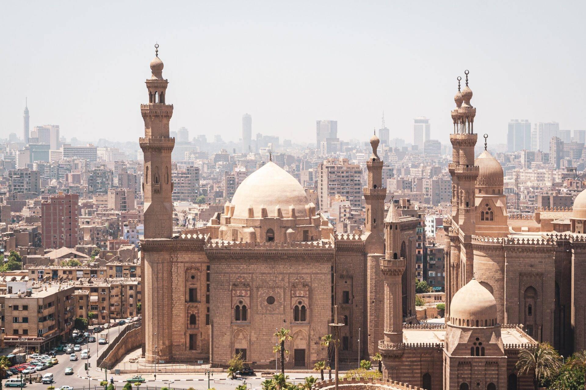 Aerial view of a historic mosque with white domes and minarets, presiding over a densely built urban neighbourhood