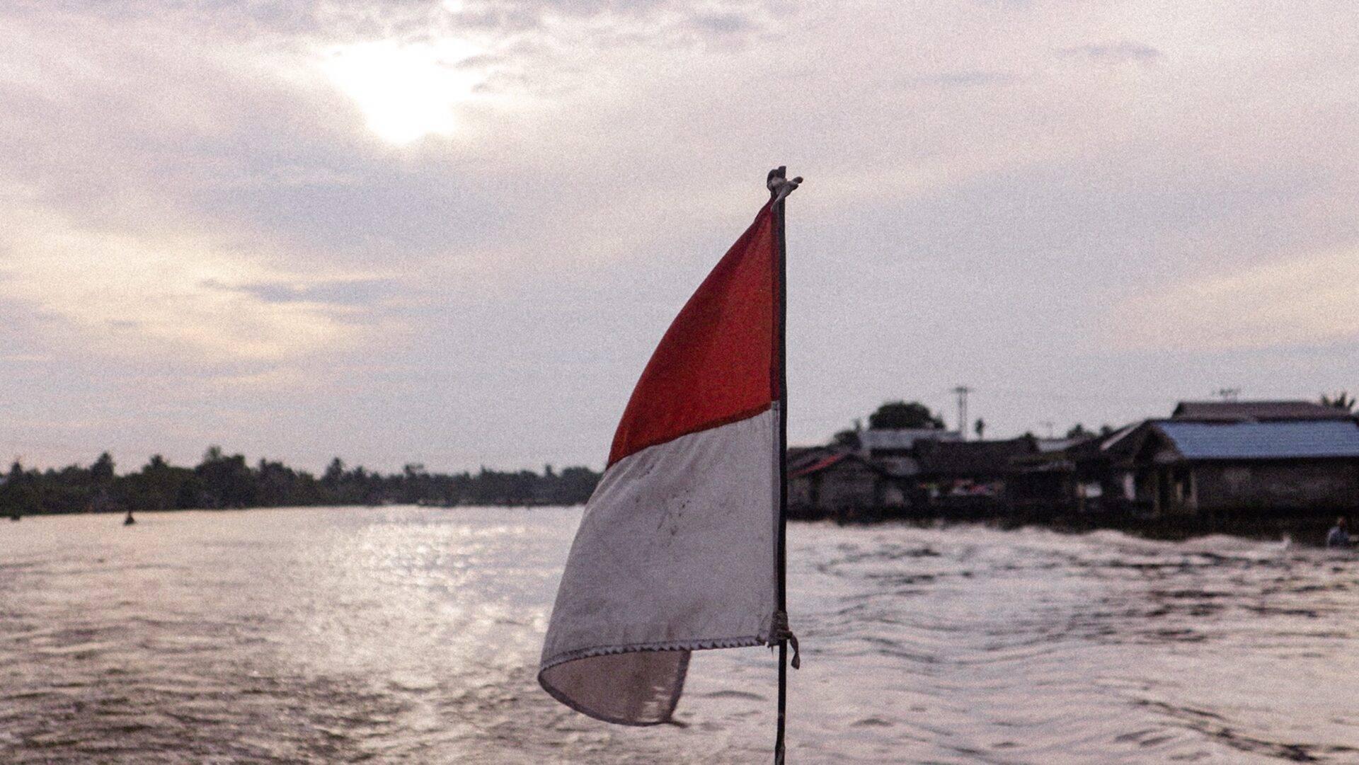 A red and white flag flying above a river with traditional houses in the background