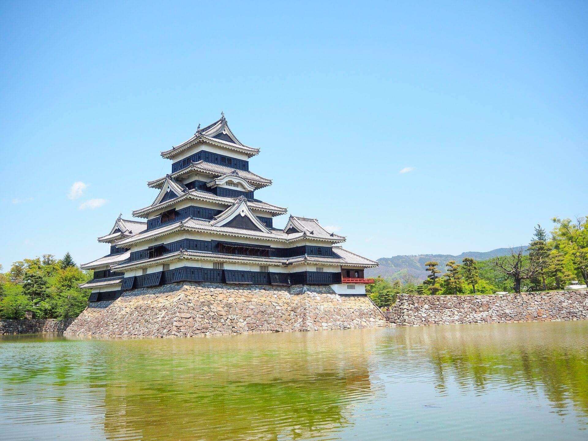A traditional Japanese castle with tiered rooflines overlooking a water-filled moat, surrounded by greenery
