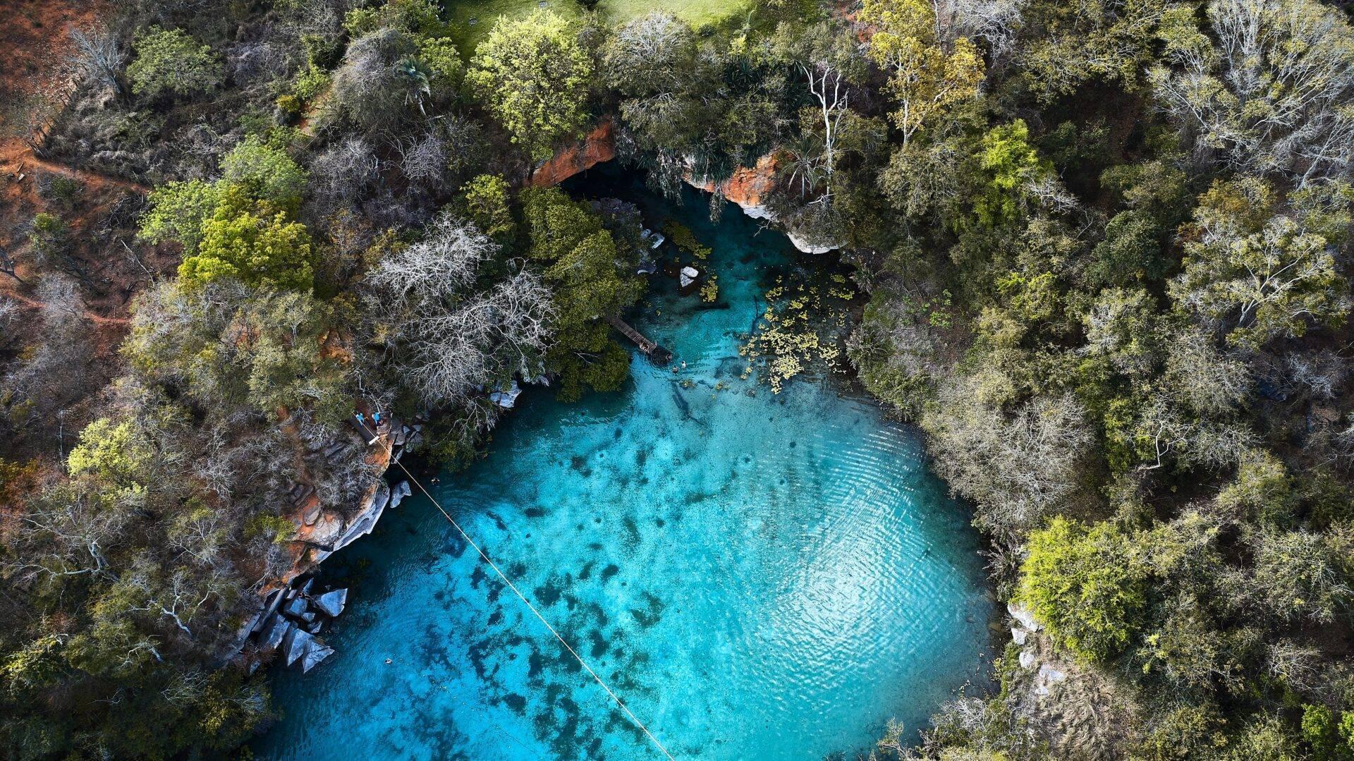 Aerial view of a turquoise pool surrounded by dense forest and rocky cliffs