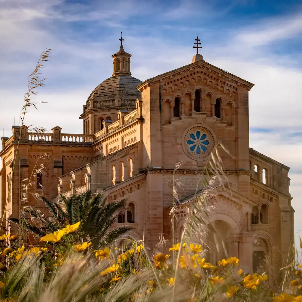 Cathédrale en pierre beige avec dôme et croix, entourée de fleurs jaunes et palmiers sous ciel bleu
