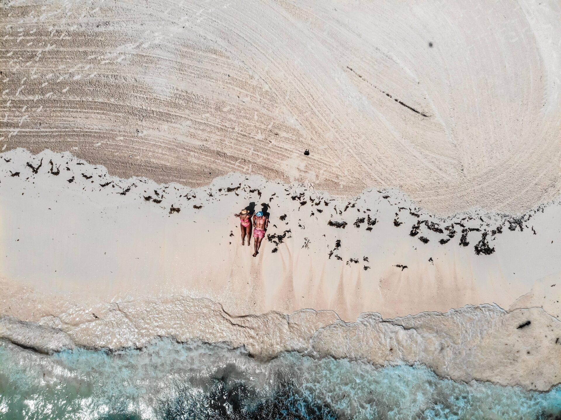 Deux personnes en maillot de bain sur une plage de sable blanc face à des vagues turquoise cristallines