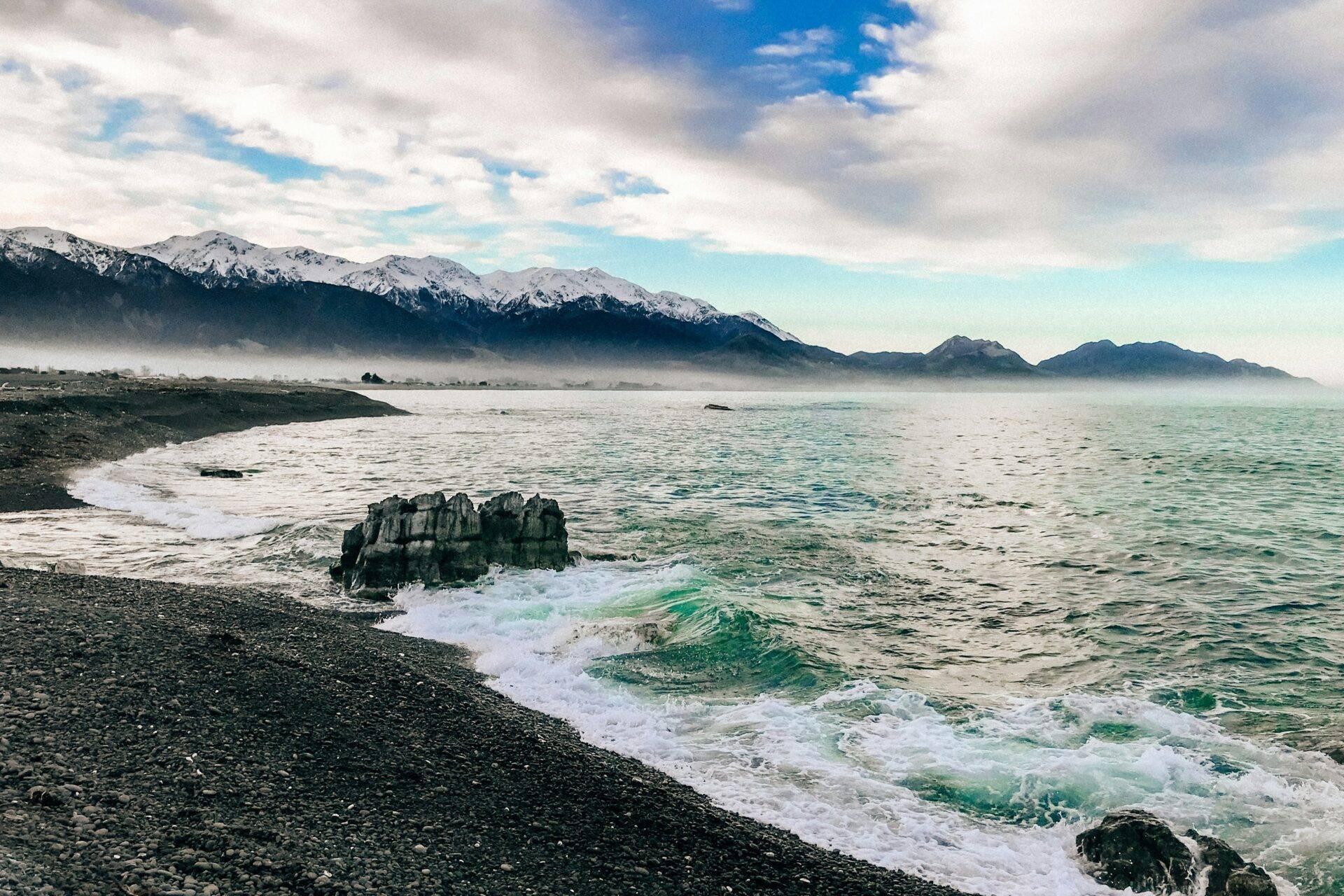 Côte rocheuse avec vagues turquoise, montagnes enneigées en arrière-plan et brume matinale sur la baie