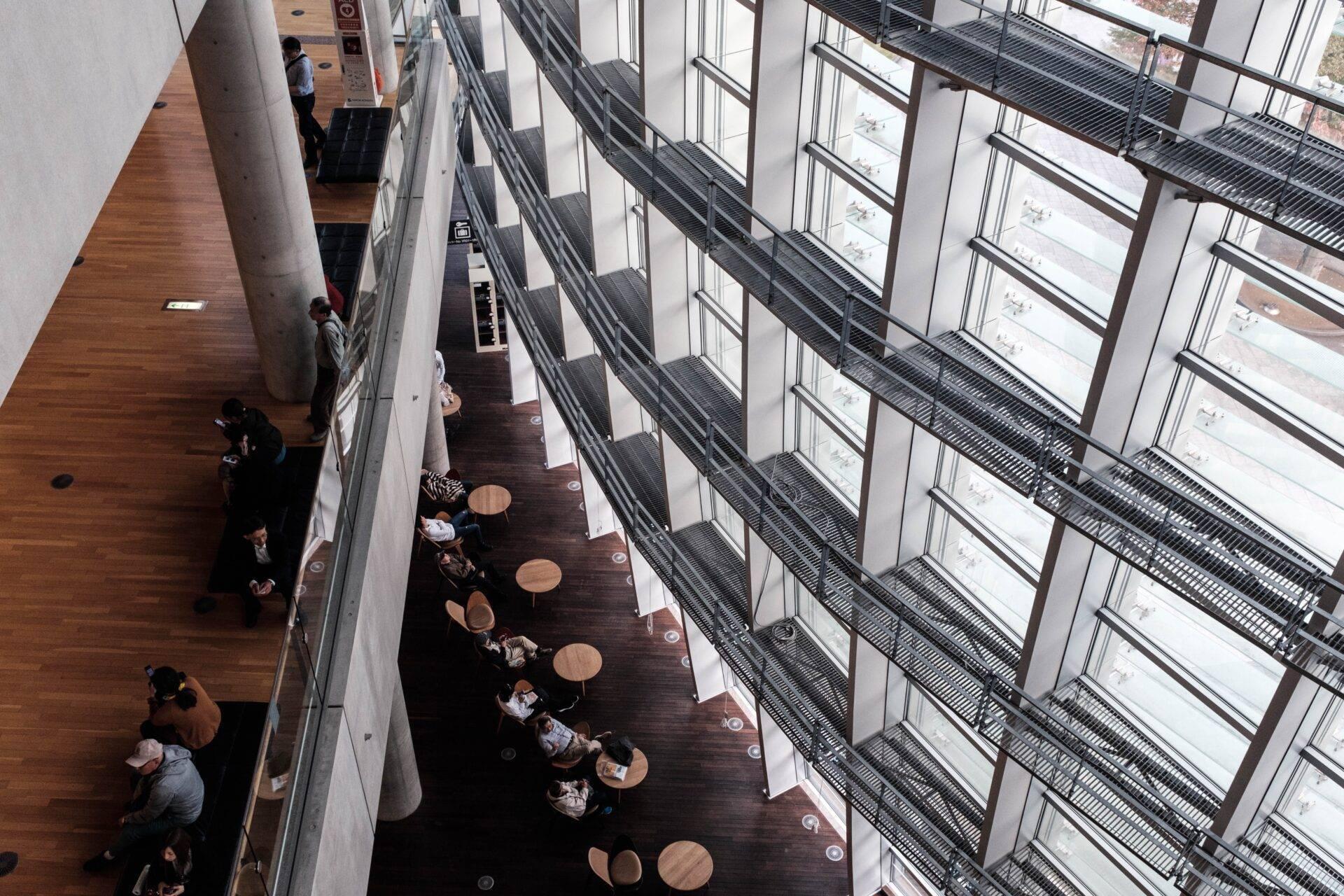 Aerial view of a modern atrium with floor-to-ceiling windows, where visitors sit at round tables along a sunlit promenade