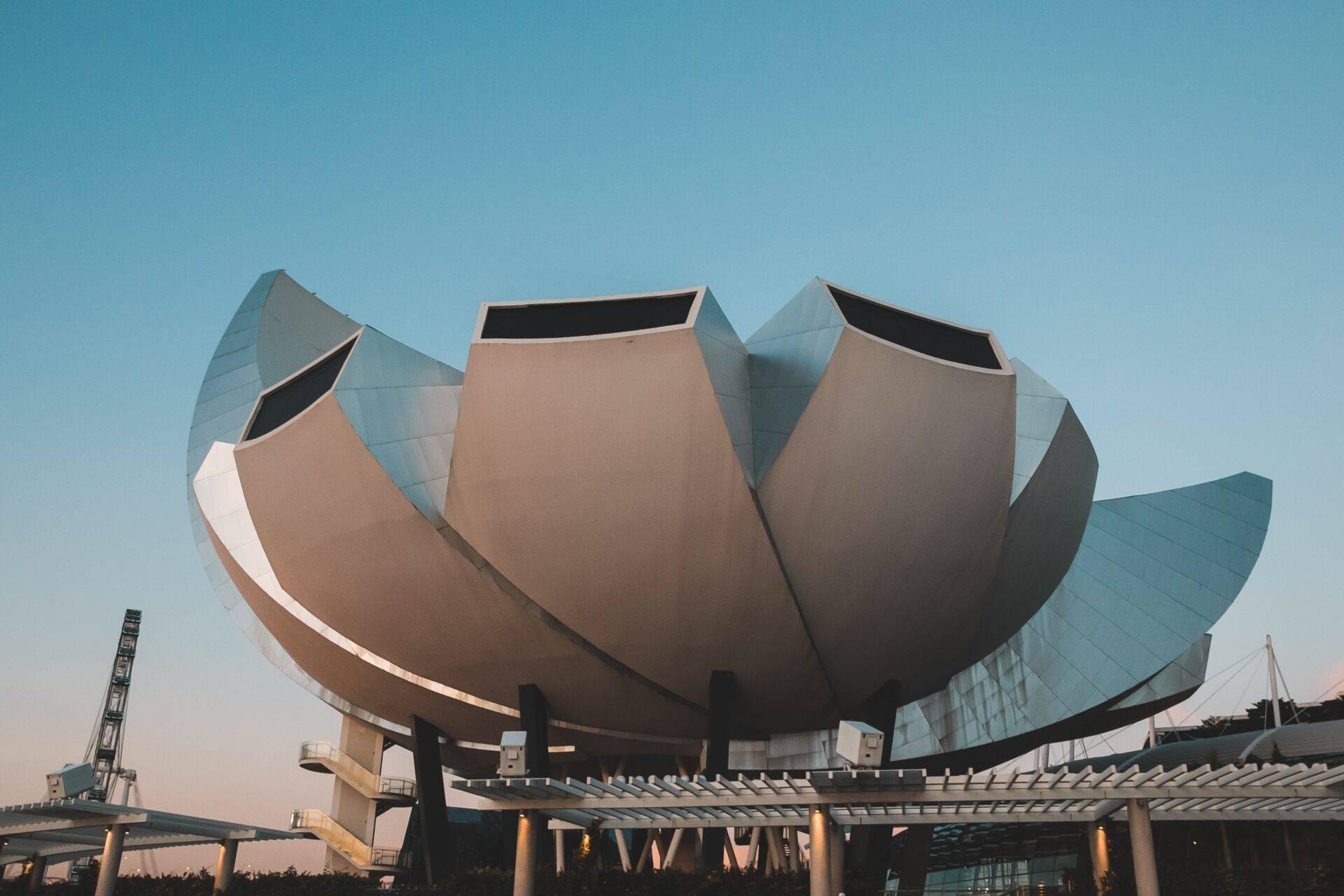 Un bâtiment architectural moderne en forme de lotus avec pétales métalliques et façade en verre sous un ciel bleu dégagé