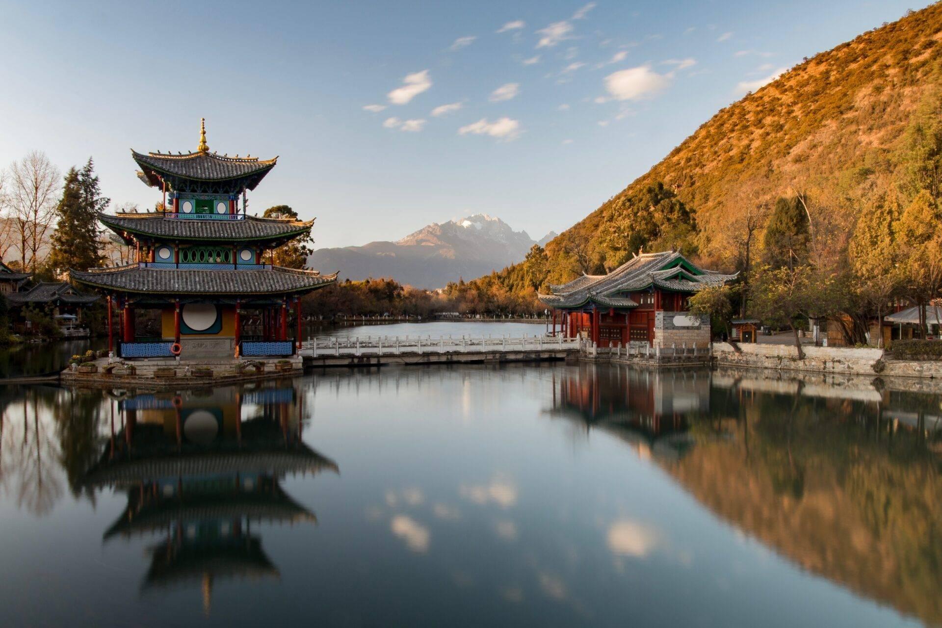 Un temple asiatique à trois étages se reflète dans un lac calme, encadré par des montagnes boisées et des pics enneigés