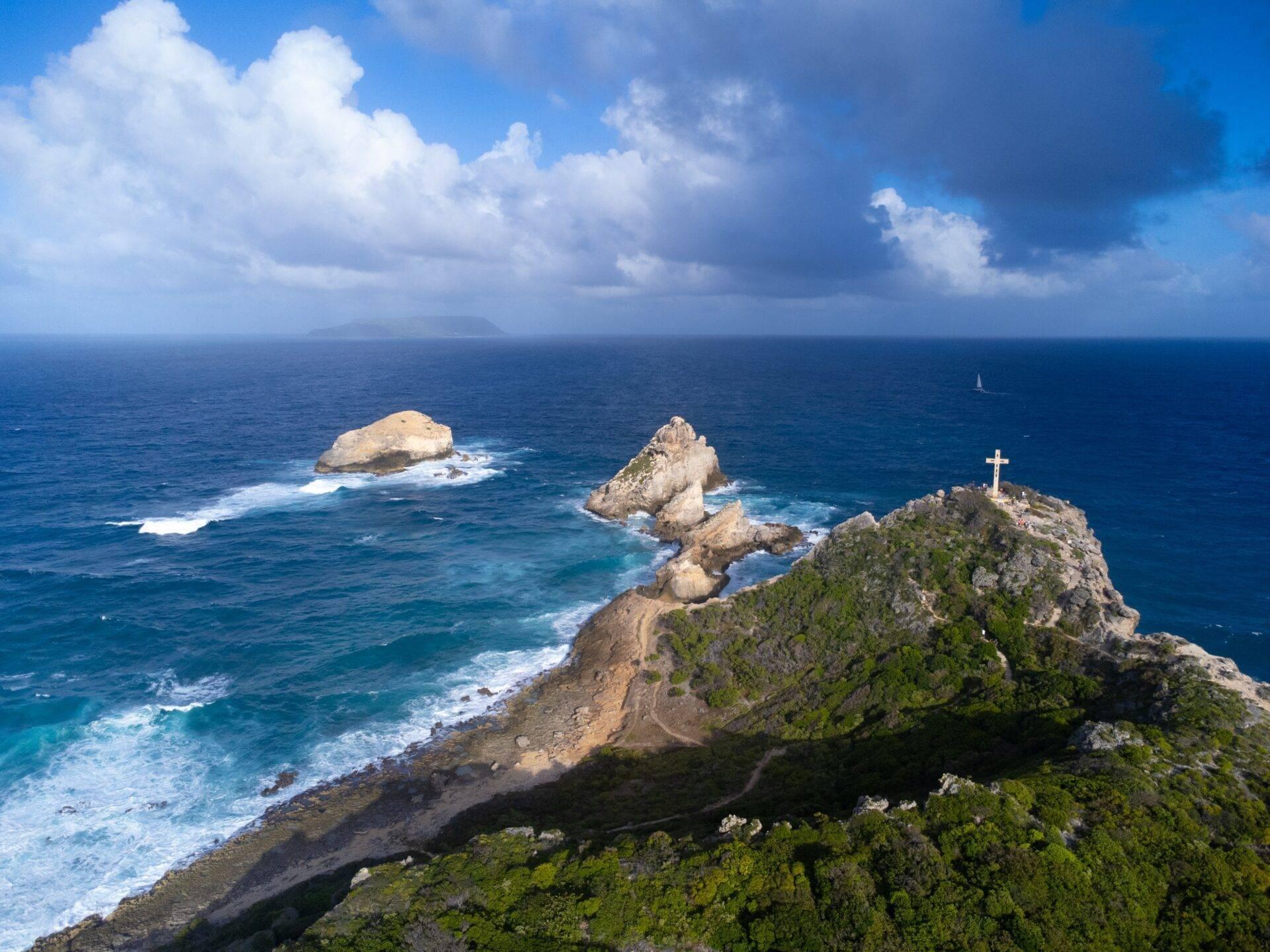 Falaise rocheuse boisée avec croix blanche surplombant l’océan turquoise et rochers isolés sous ciel nuageux