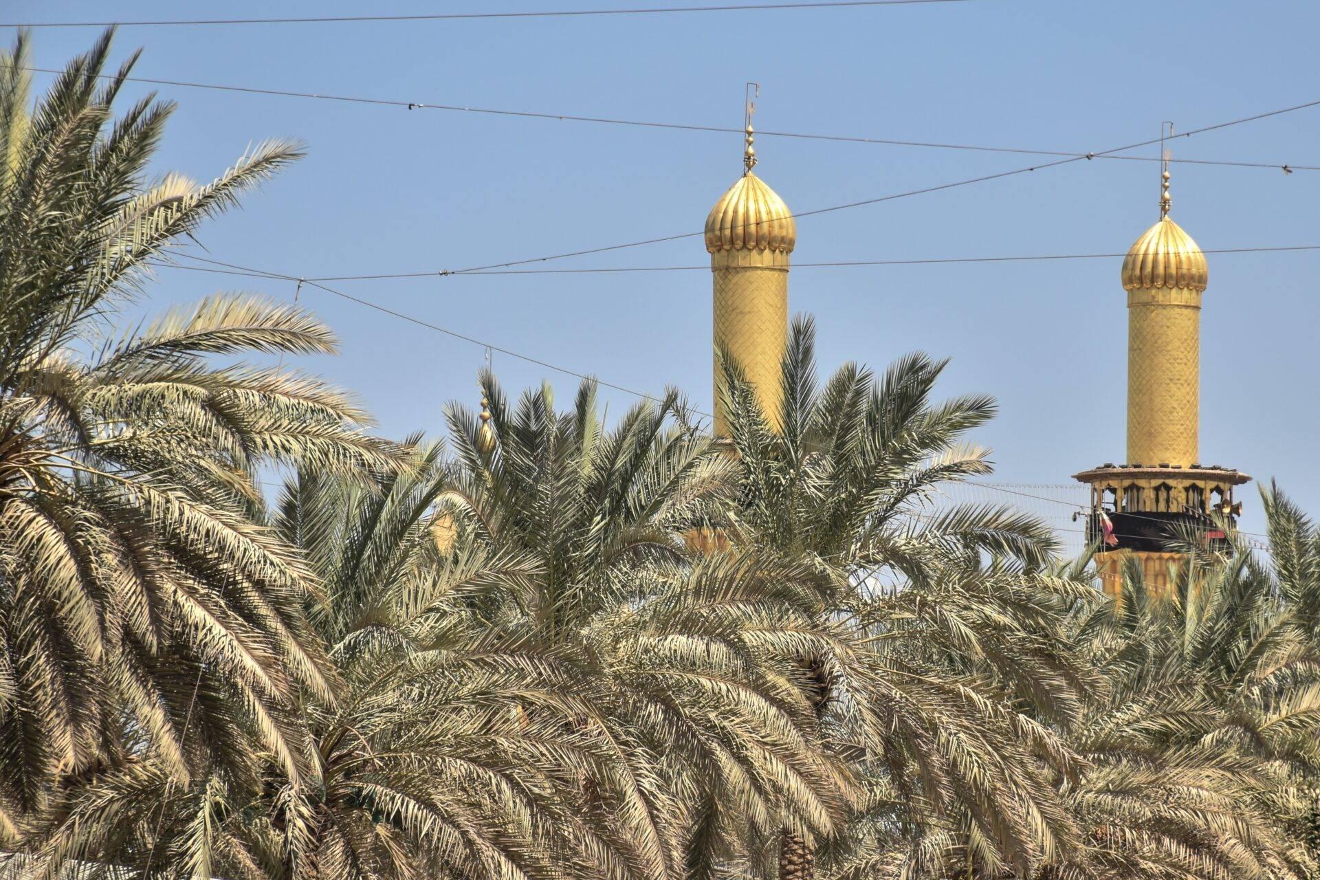 Two gilded minarets with bulbous domes rising above date palms under a blue sky