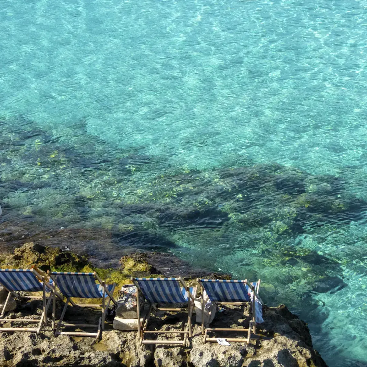 Chaises longues rayées bleu et blanc sur des rochers face à une mer cristalline méditerranéenne