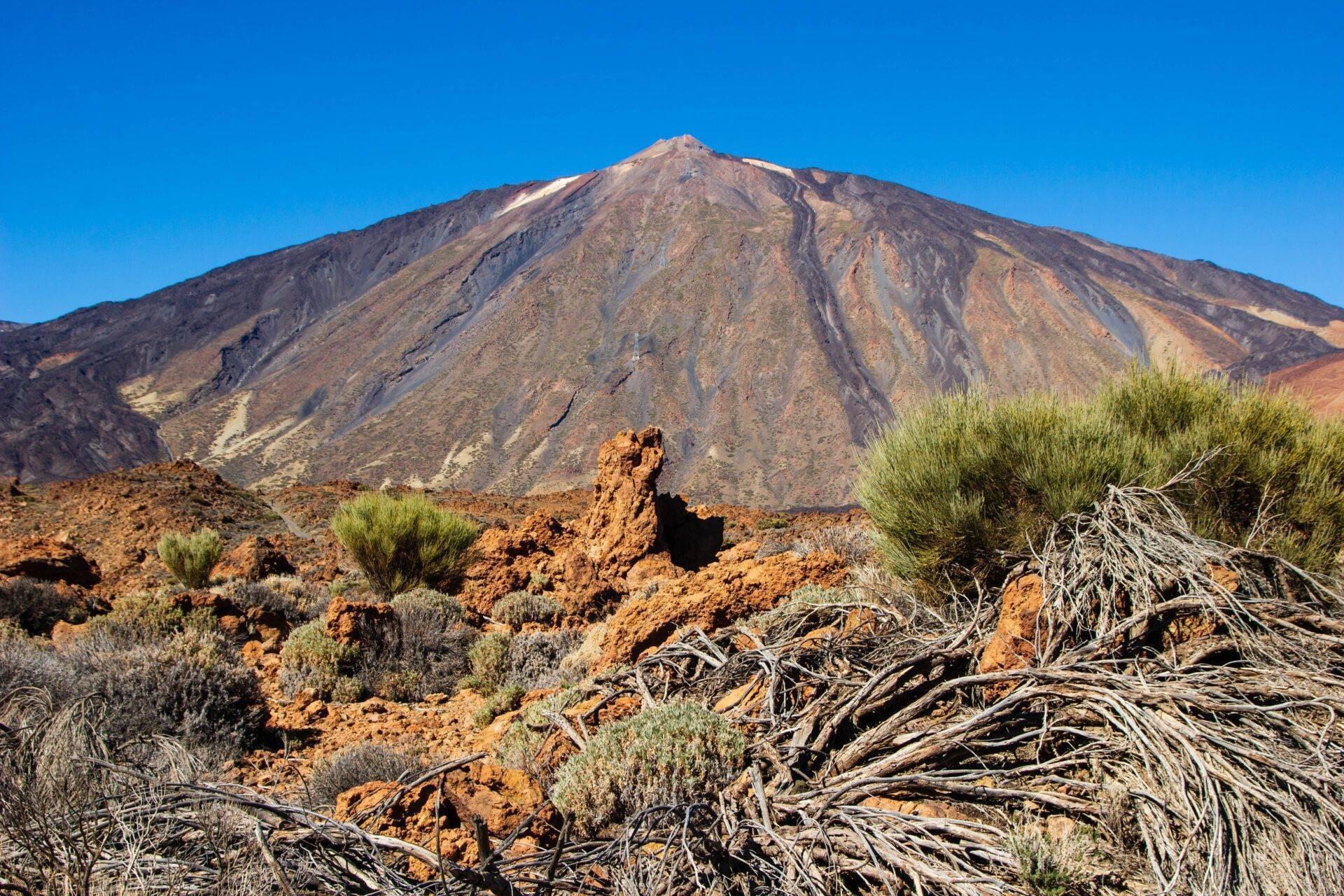 Un volcan aride aux pentes striées de minéraux colorés, entouré d’une végétation désertique clairsemée