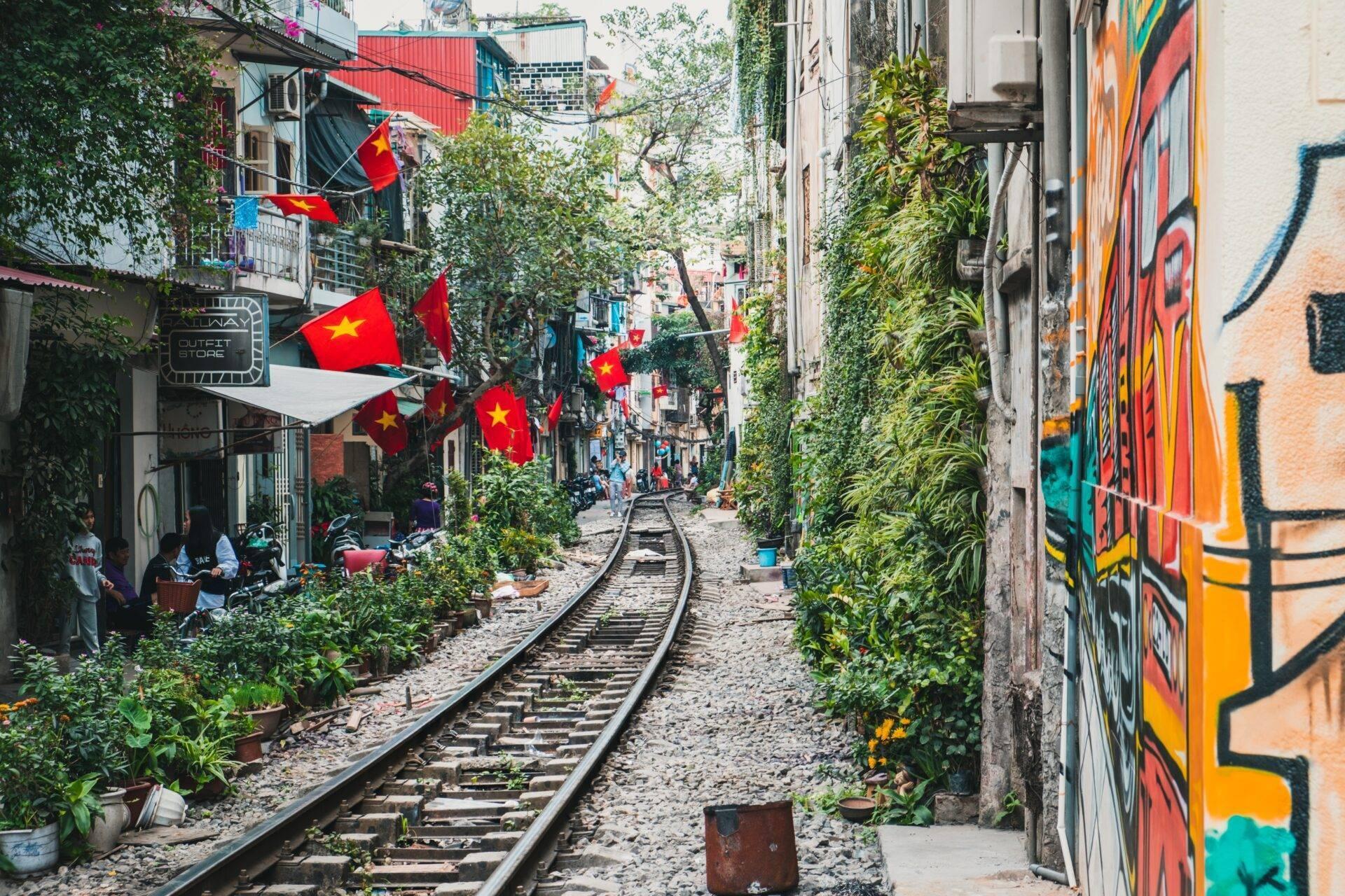 Une ruelle étroite avec voies ferrées, bâtiments colorés, végétation luxuriante et drapeaux vietnamiens rouges