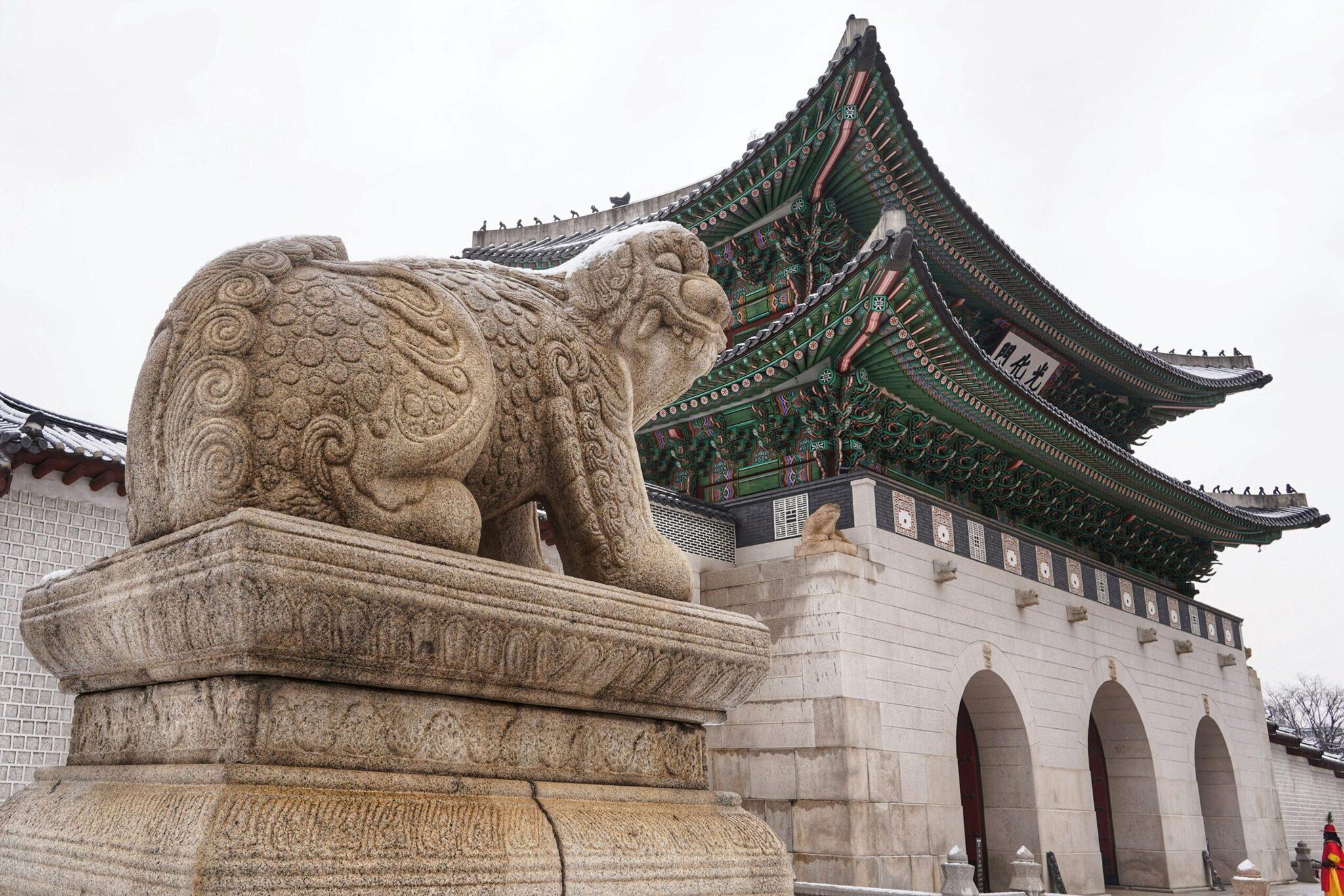 A stone lion sculpture in front of a traditional Korean pavilion with ornate green and red tiered roofs
