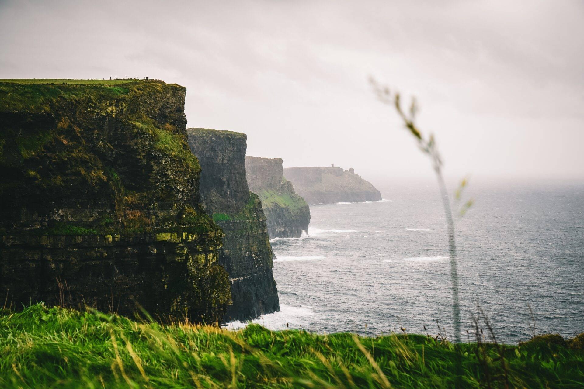 Green moss-covered coastal cliffs rising above the Atlantic Ocean under a cloudy sky