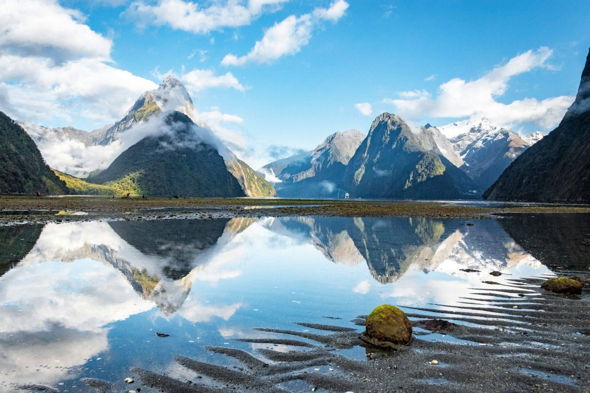Fjord montagneux avec pics enneigés se reflétant dans des eaux calmes sous un ciel bleu nuageux