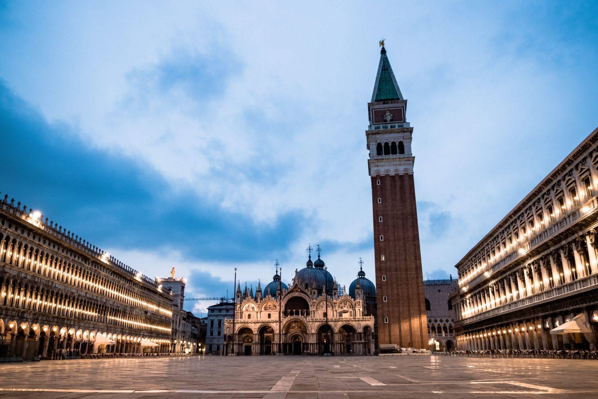 A vast paved square framed by illuminated palaces, with an ornate basilica and a red-brick campanile