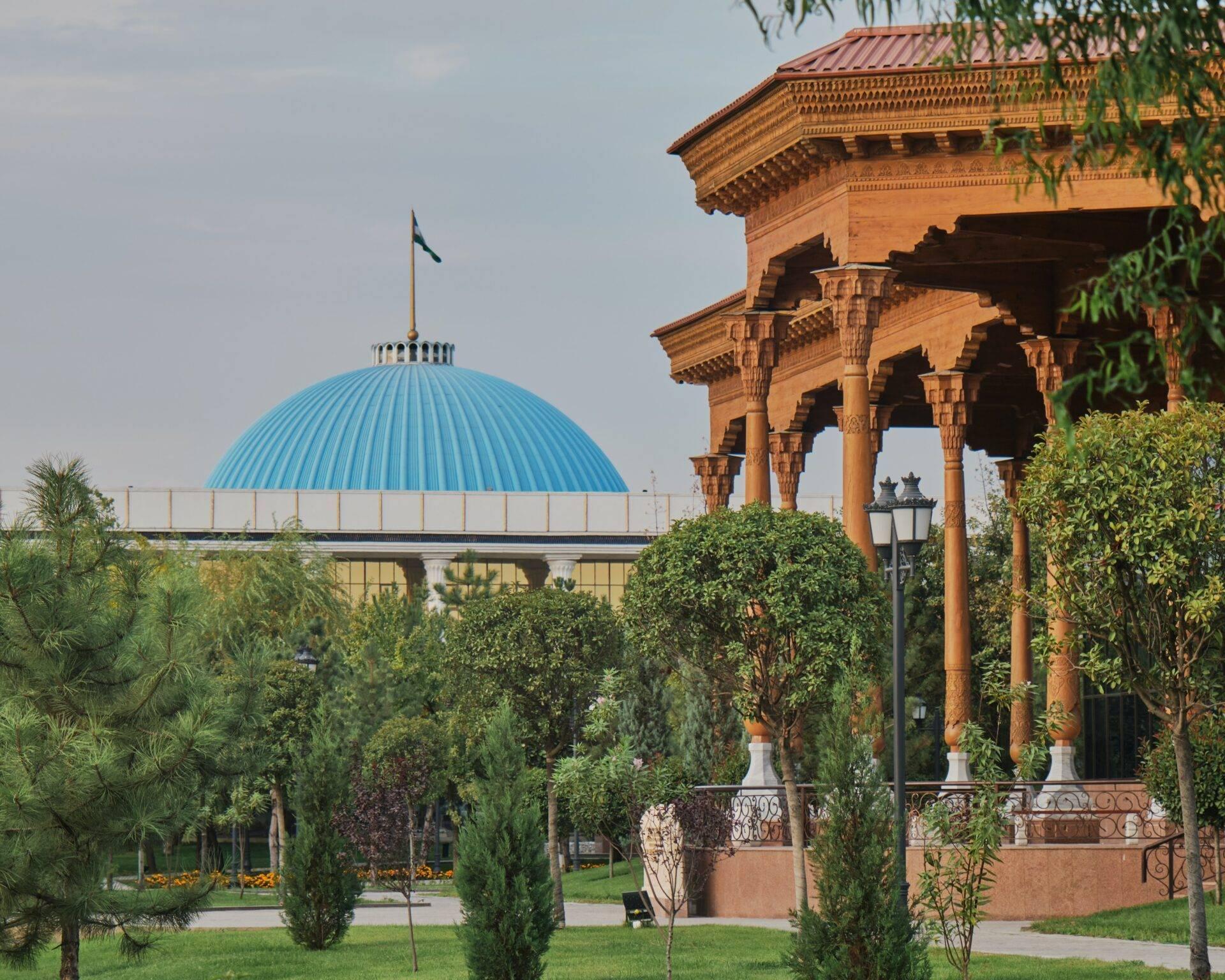 Green park with a colonnaded red-brick pavilion and a modern building topped with a turquoise blue dome
